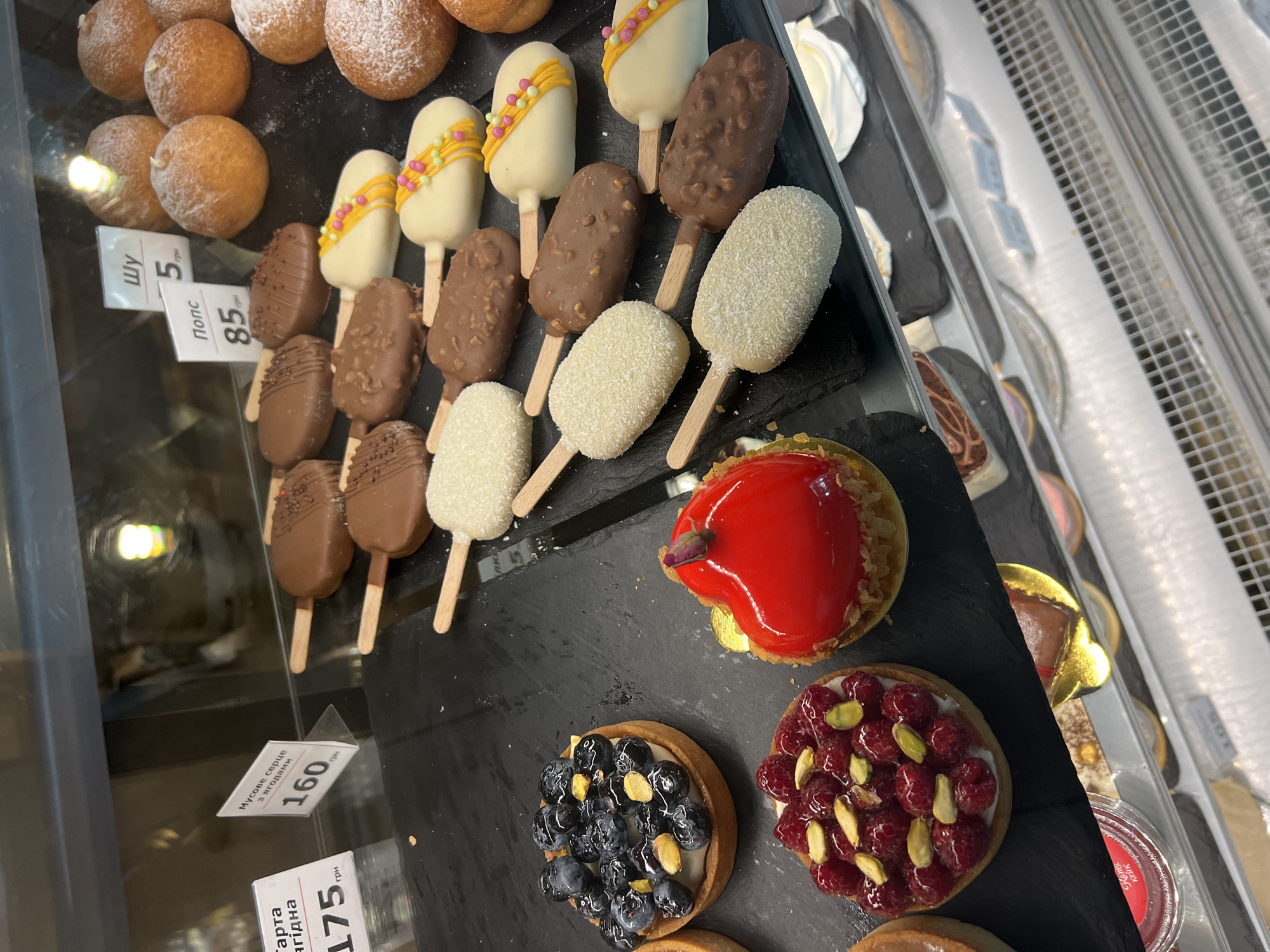 The image shows a display case in a confectionery shop, likely in Fontanka, Ukraine, filled with various desserts. In the foreground, on the left, are two small tarts: one topped with blueberries and pistachios, and another heart-shaped red glazed dessert with a small rosebud on top, also adorned with raspberries and pistachios. To the right, a selection of ice cream bar-shaped pastries are arranged on a black surface. These include some covered in chocolate and nuts, others in white chocolate with colorful sprinkles, and some coated in shredded coconut. Further back, several round, powdered doughnuts are visible. Price tags are displayed for some items, including one labeled "Мусове серце з ягодами" (Mousse heart with berries) for 160 UAH, and another for "Попс" (Pops) at 85 UAH. The display case is illuminated, highlighting the colorful and appetizing desserts. The background is slightly blurred, suggesting the shop interior.