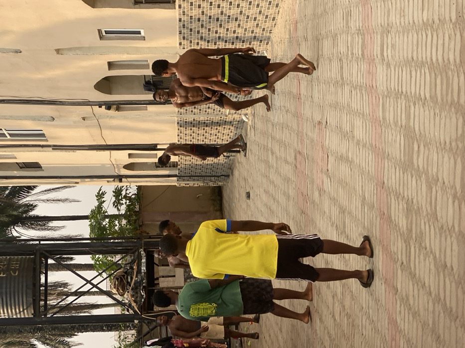 In Eziobodo, Nigeria, an outdoor scene captures seven young adult males gathered on a light-colored paved surface, featuring rectangular pavers interspersed with red lines. The setting is an alleyway or courtyard between a light-toned, multi-story building with arched ground-floor features and a brown and white brick base on the right, and a metal water tower structure with a dark water tank on the left. Several palm trees and green foliage are visible against a bright sky in the background.
The individuals are positioned across the paved area. One male, facing away from the viewer in the immediate foreground, wears a yellow short-sleeved shirt with blue trim, black shorts with white stripes, and dark flip-flops. To his left, another male wears a green t-shirt and dark shorts, standing barefoot. Further left, a shirtless male in light shorts is partially visible. Behind them, another shirtless male is seen smiling. On the right side of the frame, a shirtless male in dark shorts is smiling and facing left, displaying visible perspiration. Another shirtless male in dark shorts with a yellow-green stripe walks barefoot towards the right. Nearest the building wall on the right, a shirtless male in dark shorts stands, looking downwards, wearing dark flip-flops. The casual gathering suggests a relaxed interaction or pause in activity.