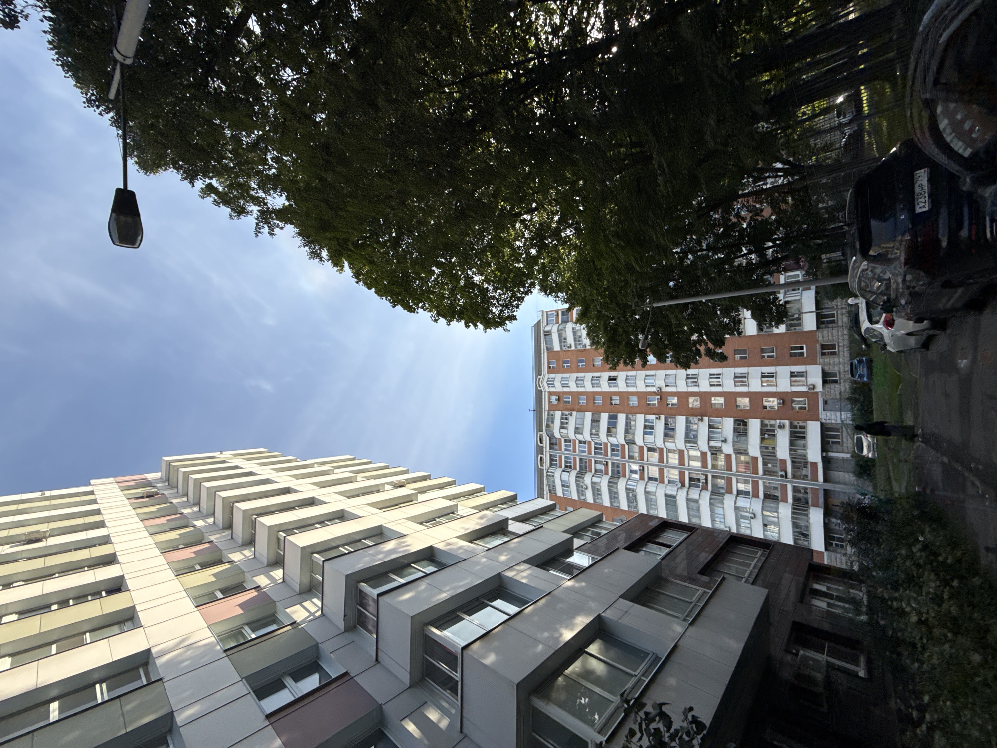 This is a ground-level, wide-angle, low-light photograph taken in a residential area of Moscow, Russia. The image captures a view of multiple apartment buildings and trees under a clear sky.
On the left, a tall, modern apartment building with a light-colored facade featuring horizontal bands of cream, pink, and gray dominates the left side of the frame, leaning significantly to the left. Adjacent to it, and closer to the foreground, is another apartment building with a darker gray facade, also appearing tall and modern, with a multitude of windows and balconies.
In the background, a much taller, older brick apartment building with numerous balconies and white window frames is visible.
A line of mature trees with dense green foliage stretches across the right side of the image, casting shadows and partially obscuring the buildings behind them. A street lamp with a dark fixture is mounted on a tall pole and is visible in the upper right quadrant of the image, partially silhouetted against the sky.
In the foreground, on a paved surface that appears to be a street or parking area, several cars are parked. The most prominent is a dark-colored SUV with a visible license plate reading "A238HP 797". Other cars, including a white sedan and a blue sedan, are also present. A single pedestrian, a person wearing dark clothing, is walking away from the camera towards the older brick building. The overall lighting suggests it might be late afternoon or early evening, with sunlight filtering through the trees. The environment appears to be a typical urban residential block with high-rise buildings and interspersed green spaces.