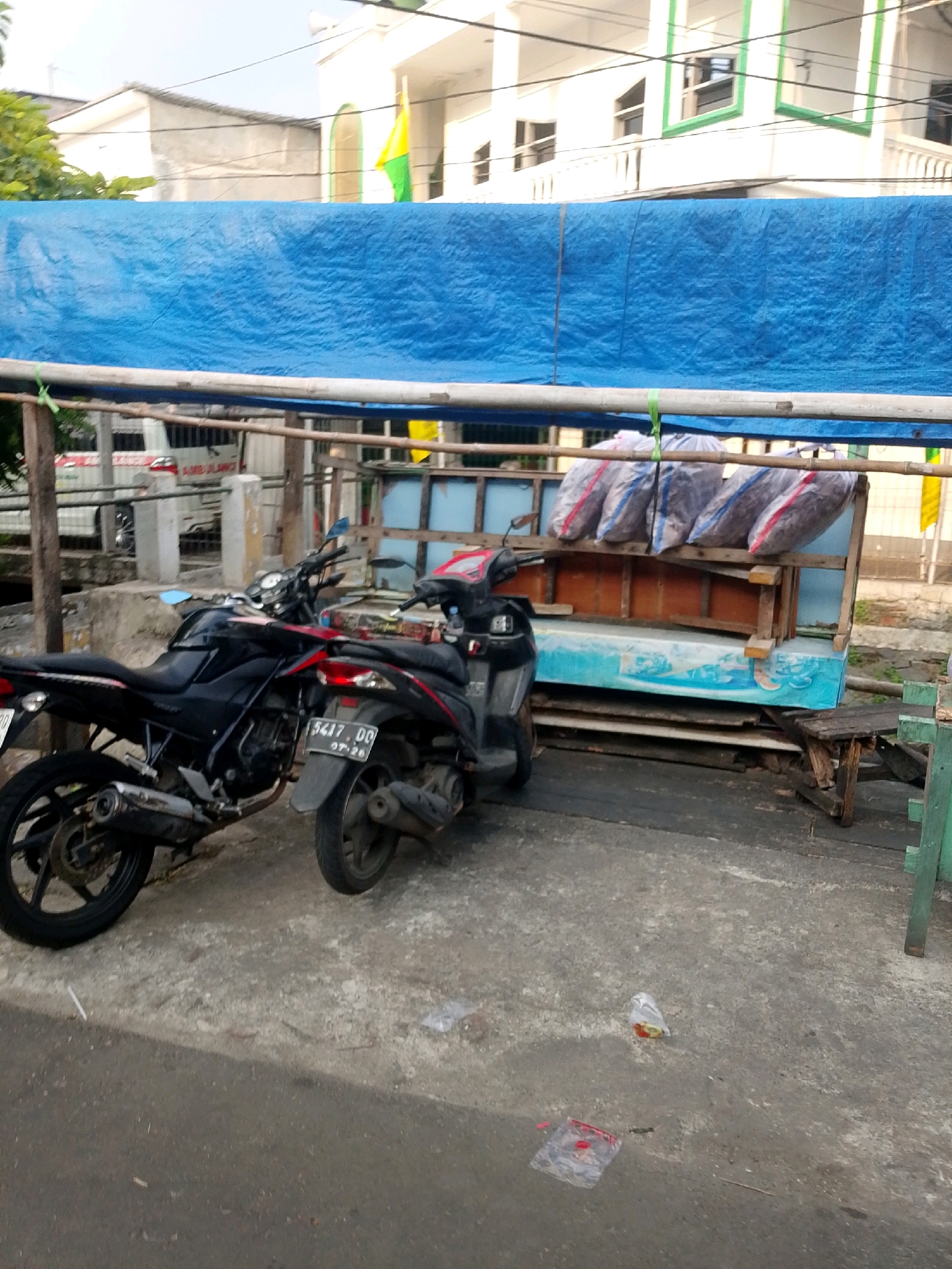 Two motorcycles are parked next to a roadside stall covered by a blue tarp in East Jakarta, Indonesia. The stall is constructed from wood and has a light blue surface at the bottom, with several bags of goods stacked on a wooden shelf above. In the background, a white building with green trim and a yellow and green flag are visible. The ground is paved with concrete and asphalt. There are no people visible in the image.