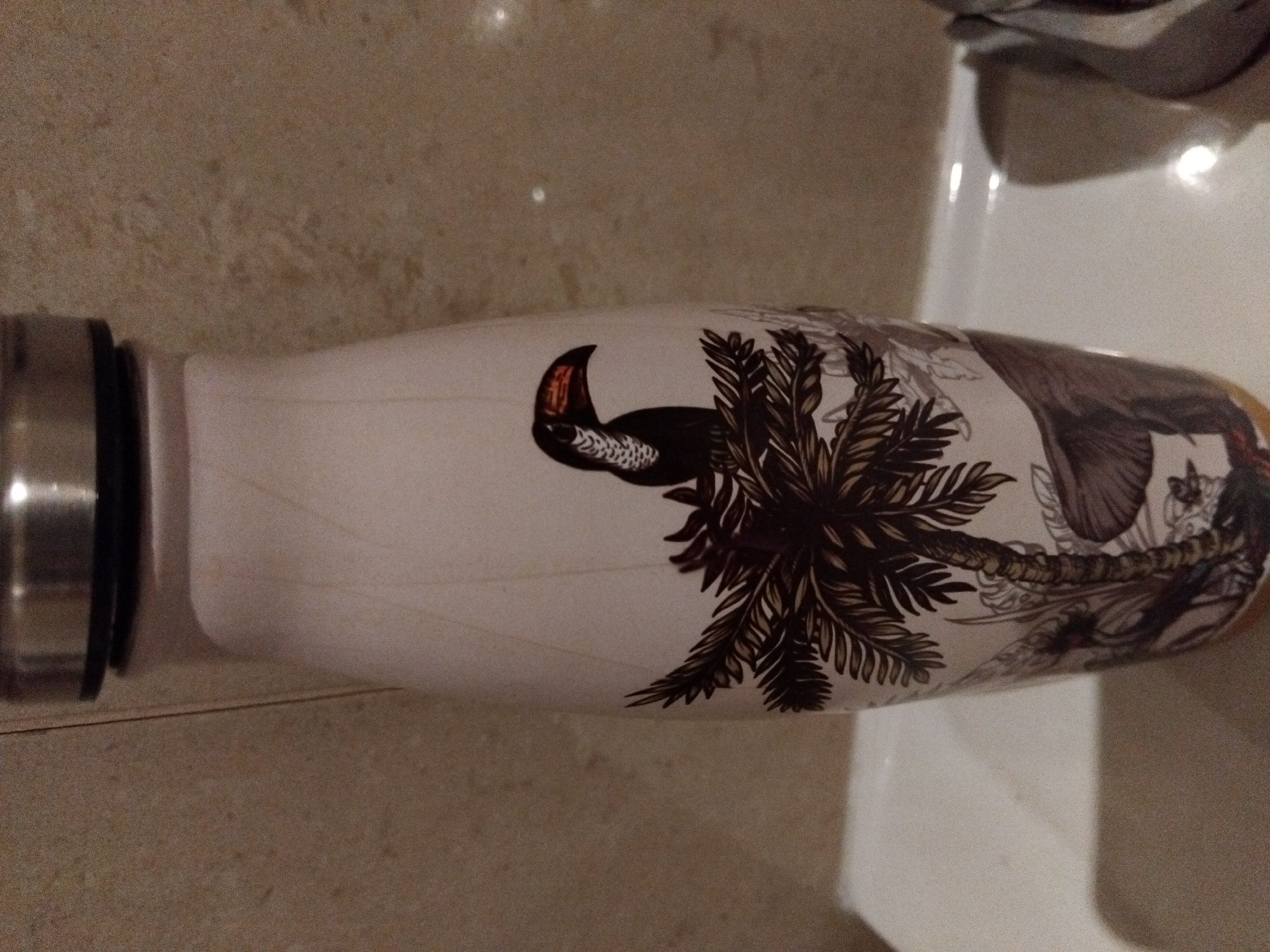 A close-up, slightly angled shot shows the upper portion of a white, cylindrical water bottle with a metallic silver lid. The bottle is decorated with a detailed, stylized illustration. On the visible side of the bottle, there is a depiction of a toucan with a black body, white chest markings, and an orange and black beak, perched on a palm tree frond. The palm tree itself is rendered in dark brown tones, with its fronds extending downwards. Beneath the palm tree, there are other tropical foliage elements and what appears to be the outline of an elephant's head. The background is a textured, light brown wall, possibly tiled, that appears to be in a bathroom setting. The bottle rests on a white surface, possibly a sink or countertop. The lighting is dim and artificial. The scene is devoid of any people or discernible human interaction. There is no information to suggest this image is located in Vila Nova de Famalicão, Portugal.