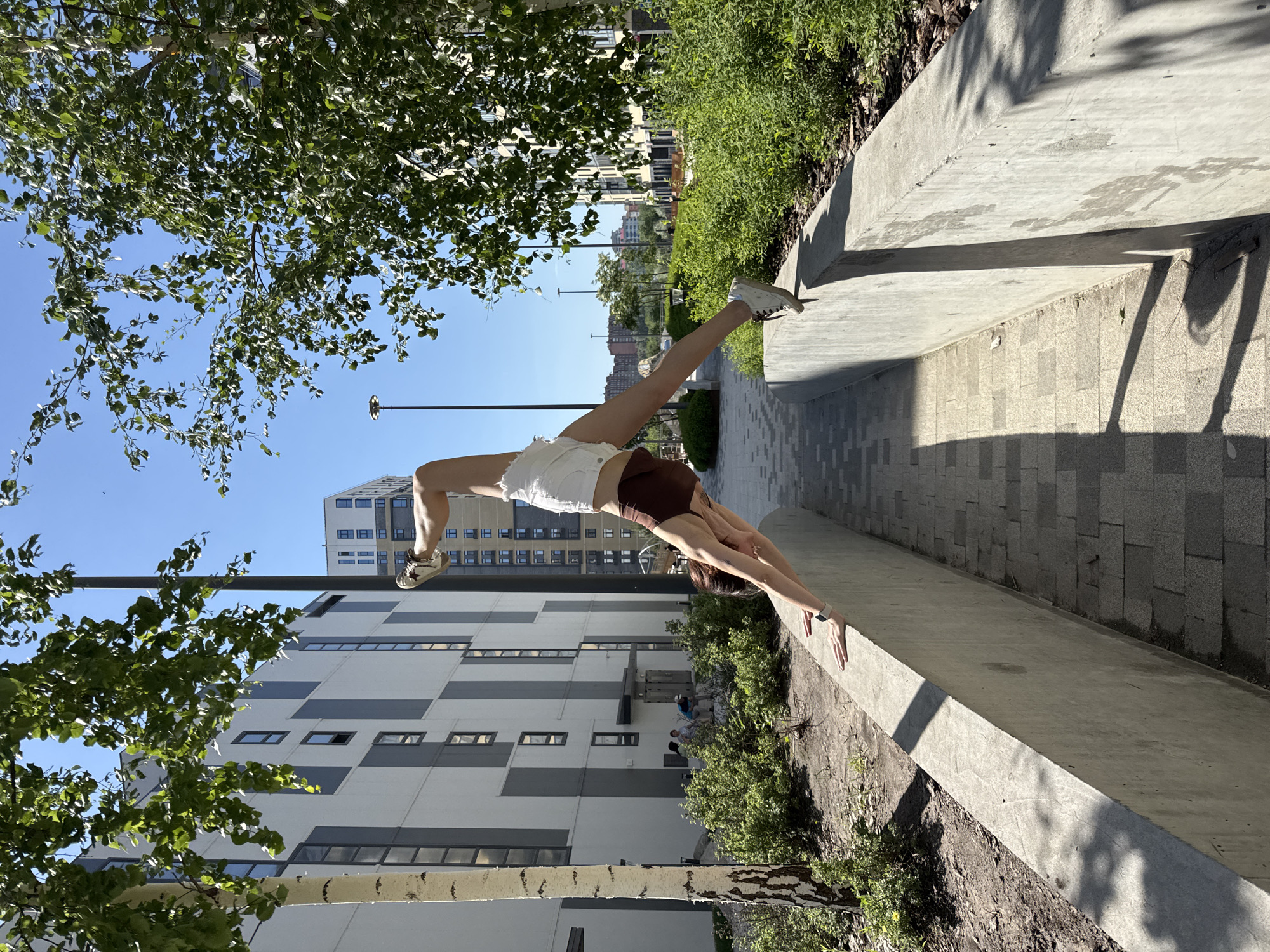 A woman is performing a backbend on a concrete ledge in an urban environment. She is wearing a brown tank top and white shorts, and white sneakers. Her feet are in the air. The background includes a modern building with gray and white paneling, greenery, and a clear blue sky. Part of a birch tree is visible with green leaves. The scene appears to be in Tyumen, Russia, based on architectural styles and urban design elements common in the city. The ledge is situated next to a paved walkway.