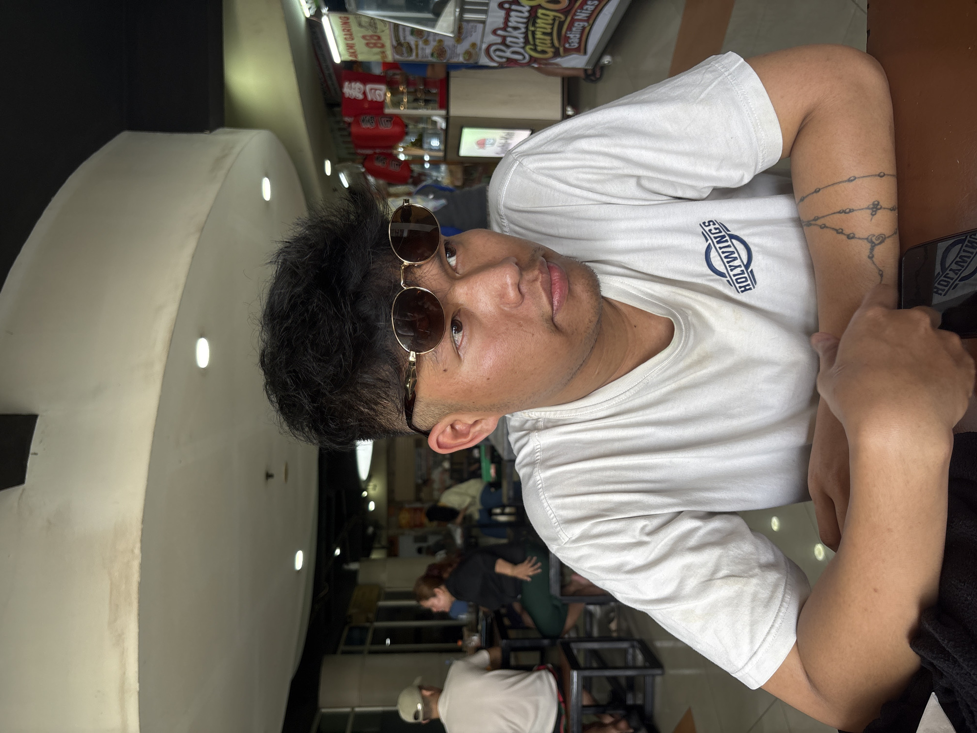 A young man wearing sunglasses and a white t-shirt with the "Holywings" logo is seated at a table in an indoor food court. He has dark, spiky hair and a tattoo on his left forearm. In the background, other patrons are visible, and stalls with food signs are set up. One sign reads "Bakmi Garing Gading Nias." The environment appears to be brightly lit and moderately busy. This scene is consistent with a food court in a shopping mall or commercial area in North Jakarta, Indonesia, possibly in a Holywings establishment.