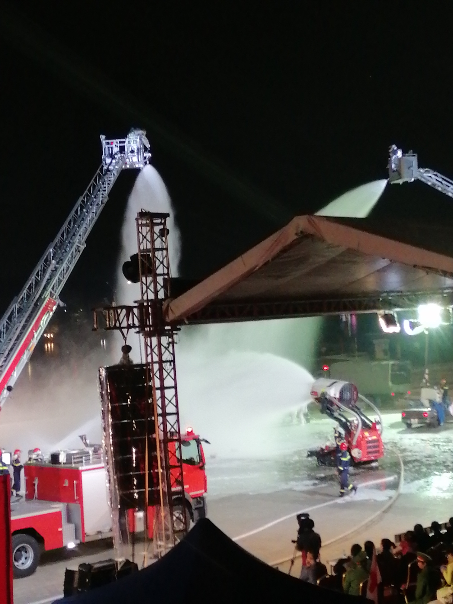 The image captures a nighttime scene in Da Lat, Vietnam, where a group of firefighters are actively engaged in a water display, likely part of a public event. Two large fire trucks are prominently featured. One is a red and white truck on the left, with its ladder extended upwards. Another red fire truck is positioned towards the right, and it appears to be equipped with a rotating water cannon that is actively spraying a large volume of water into the air, creating a misty effect. This water cannon is mounted on a red vehicle.
In the background, a stage or covered structure is visible, illuminated by bright lights. Several figures, presumably firefighters in uniform, are positioned around the equipment, operating the water spray. One firefighter is seen wearing a blue and yellow uniform, while others are in darker attire. The water spray creates a dynamic visual effect, catching the light and creating a luminous mist.
In the foreground, a crowd of people is gathered, observing the demonstration. Many are silhouetted against the bright lights, but some are discernible, suggesting a festive or celebratory atmosphere. The overall setting appears to be an open area, possibly a public square or event ground, given the presence of the crowd and stage. The event is taking place at night, indicated by the dark sky and artificial lighting. There are no discernible emotions, but the scene suggests a coordinated and perhaps entertaining demonstration. No visible text is present in the image.