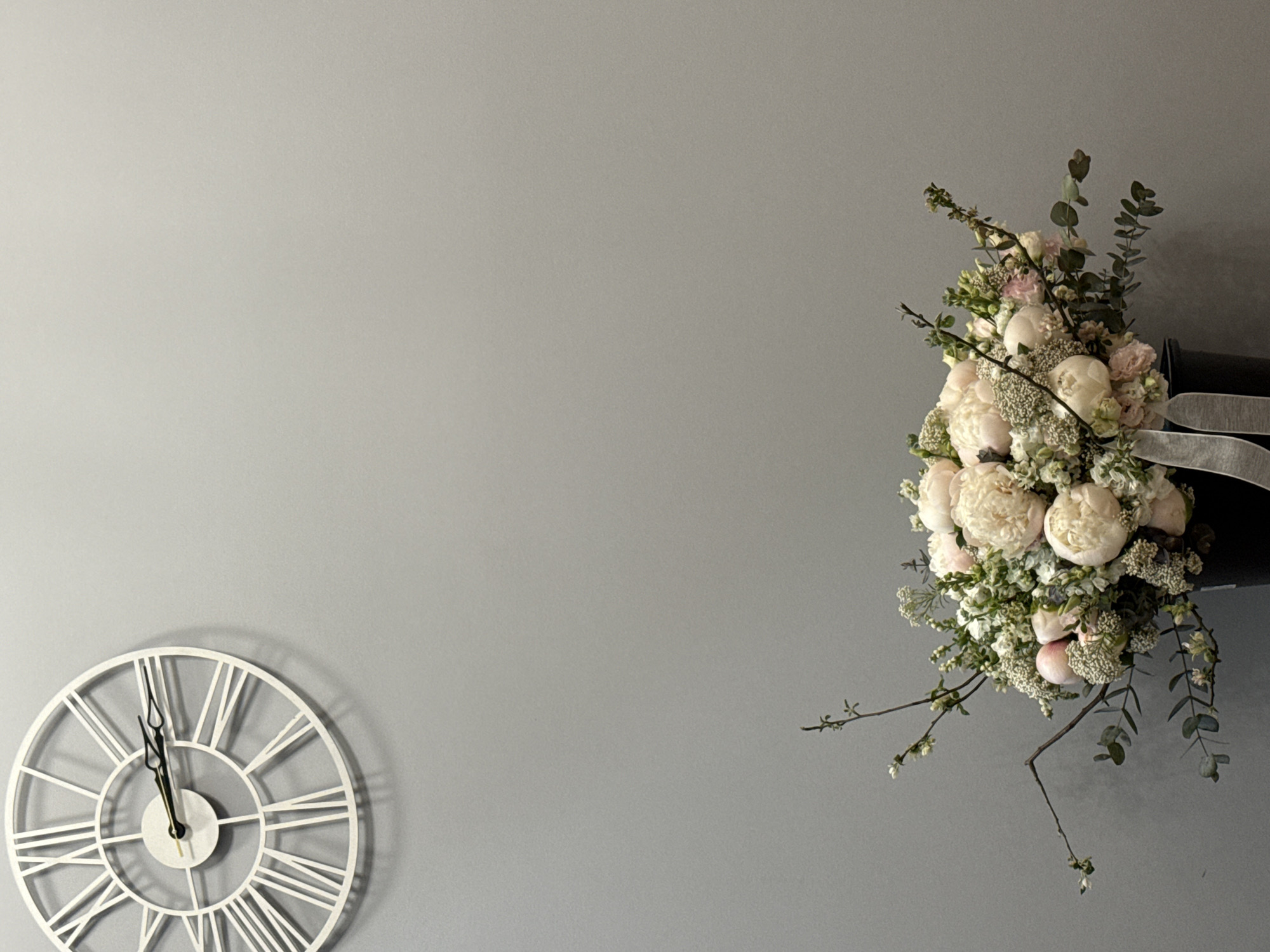 The image shows a wall with a white, decorative clock on the lower left side and a floral arrangement on the upper right side. The clock face indicates a time of approximately 11:57. The floral arrangement appears to be peonies and greenery in a dark container. The wall is a neutral grey color. No people are visible. The location in Moscow, Russia, is not identifiable from the visual information.