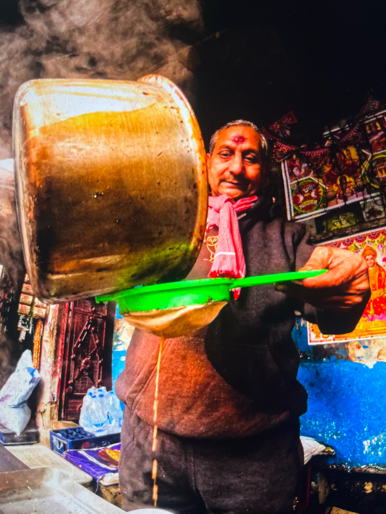 An adult male with a red tilak on his forehead and a pink scarf around his neck is pouring a golden liquid from a large metal pot into a green strainer. The liquid is steaming. He is wearing a brown hooded sweatshirt and dark pants. The background consists of a vibrant blue wall with colorful religious posters and some white sacks and plastic bottles. This scene appears to be from a street vendor stall in Gola Gokarannath, India, likely preparing a beverage such as chai.