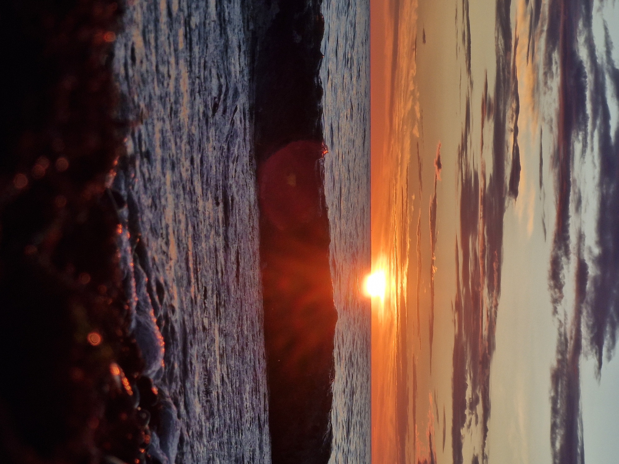 The image depicts a sunset over the Baltic Sea. The sun, a bright orb, is positioned near the horizon, casting a warm orange glow across the sky and the water. Clouds are scattered across the sky, reflecting the colors of the sunset. A wave is breaking in the foreground, with foam and spray visible. The foreground is dark and out of focus, with some bokeh effects of light. The scene appears to be a natural landscape, likely a beach or coastline. The image does not contain any people, objects, or actions, and it is not possible to determine the specific location in Klaipėda, Lithuania from the visual information alone.