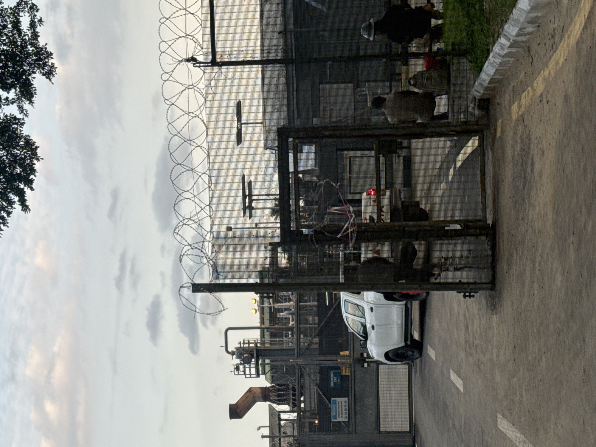 The image depicts an industrial facility in Igwuruta, Nigeria, secured by a tall metal fence topped with barbed wire. A white pickup truck is parked near the entrance gate. Industrial structures with pipes, tanks, and machinery are visible within the compound. Overhead, the sky is overcast. Some workers are visible within the compound, near the gate. The setting appears to be a roadside, with a curb and pavement markings visible.