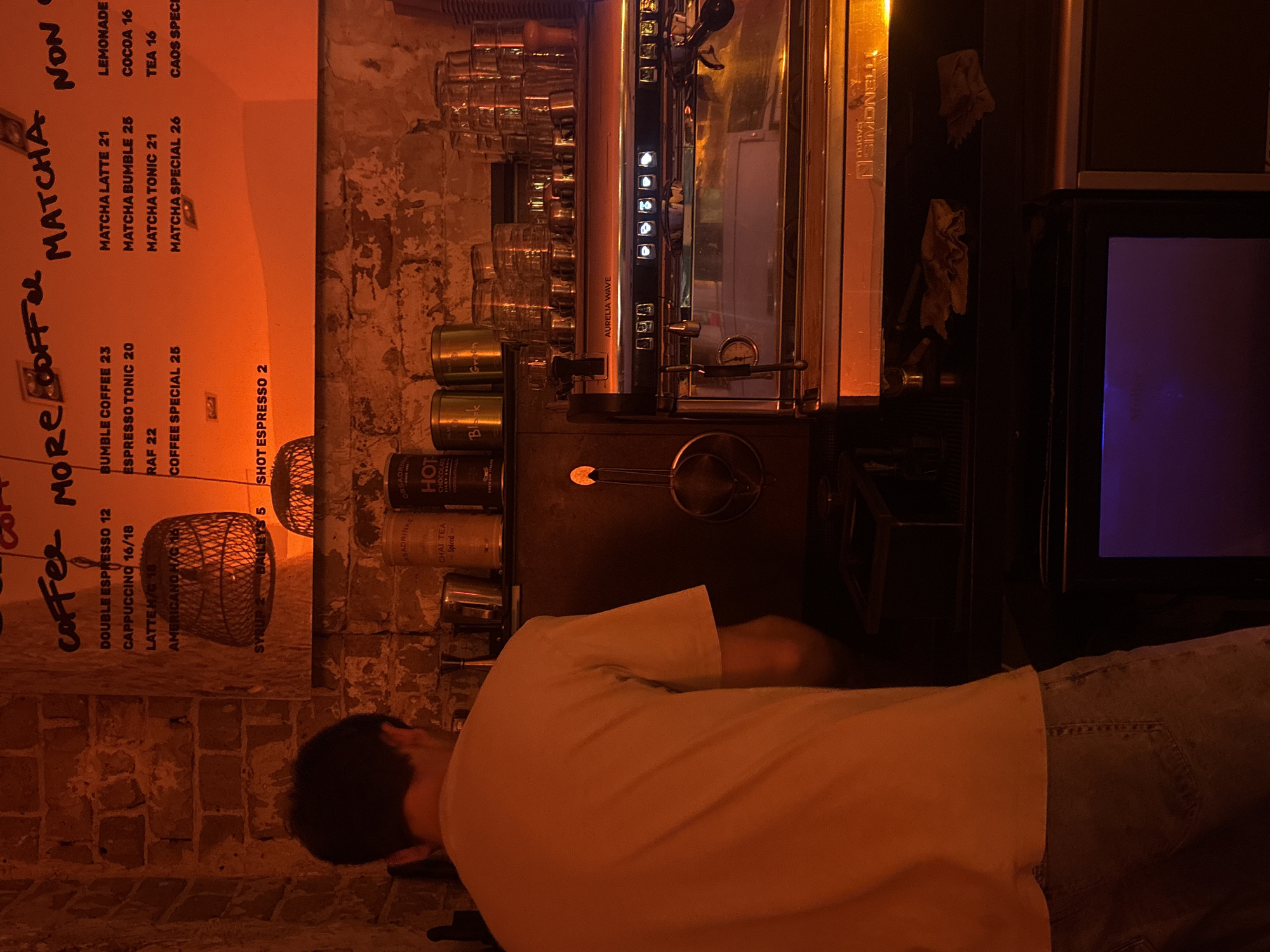 The image captures a dimly lit interior scene, possibly a coffee shop or bar, with a warm, orange glow emanating from overhead lighting and a large menu board. A person, seen from behind, is wearing a light-colored t-shirt and jeans and appears to be standing in front of a coffee machine. The coffee machine is a prominent stainless steel appliance with various buttons and indicators. On shelves above the machine, there are stacked glasses, cans labeled with text like "CHAI TEA" and "HOT CHOCOLATE," and other bar-related items. The background consists of a brick wall. The menu board displays various coffee and drink options with prices, including "DOUBLE ESPRESSO 12," "CAPPUCCINO 16/18," "MATCHA LATTE 21," and "NON CAOS 16." While the image's visual cues do not explicitly place it in Tel-Aviv, Israel, the presence of Hebrew script on some items, such as the cans and the menu board's organization, suggests a strong likelihood of this location. Specifically, the menu items and pricing structure are consistent with establishments found in Tel-Aviv.