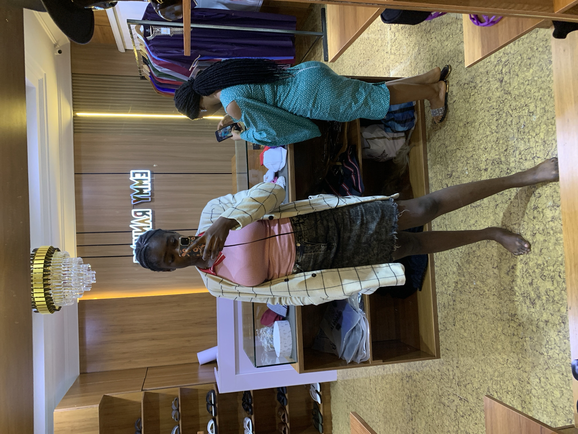 In Gura Top, Nigeria, a young Black woman stands barefoot in a store, taking a selfie. She wears a light-colored plaid jacket over a pink and white striped shirt and a dark denim skirt. To her right, another Black woman, wearing a teal patterned dress, is looking at her phone. The store environment features wooden shelves filled with clothing and merchandise. A neon sign with blurred text, likely a store name, is visible on the wall. A modern chandelier hangs from the ceiling. The floor has a textured yellow pattern.