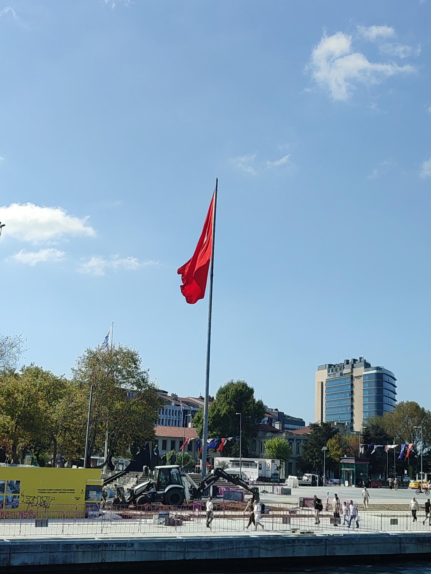 The image depicts a wide, outdoor urban scene on a clear, sunny day with a bright blue sky and scattered white clouds. A prominent large Turkish flag, red with a white star and crescent, flies high on a tall pole to the left of the frame. In the background, there is a modern, glass-fronted building with multiple stories. Trees with green foliage are visible throughout the middle ground, interspersed with older, more traditional buildings and some construction elements.
In the foreground, a body of water, likely a harbor or bay, takes up the bottom portion of the image. A paved promenade or walkway runs along the edge of the water, separated by a metal barrier. On this walkway, several people are casually walking in various directions. To the left of the walkway, there is a construction site with heavy machinery, including a backhoe loader. A large yellow construction barrier with text and graffiti is also present. Flags of various colors are displayed on poles in the middle ground, and a yellow vehicle, possibly a taxi, is seen on the right side of the promenade.
Based on the presence of the large Turkish flag and the architecture, the scene is likely in Kadıköy, Turkey. The overall impression is of a bustling public space undergoing some form of development or renovation.