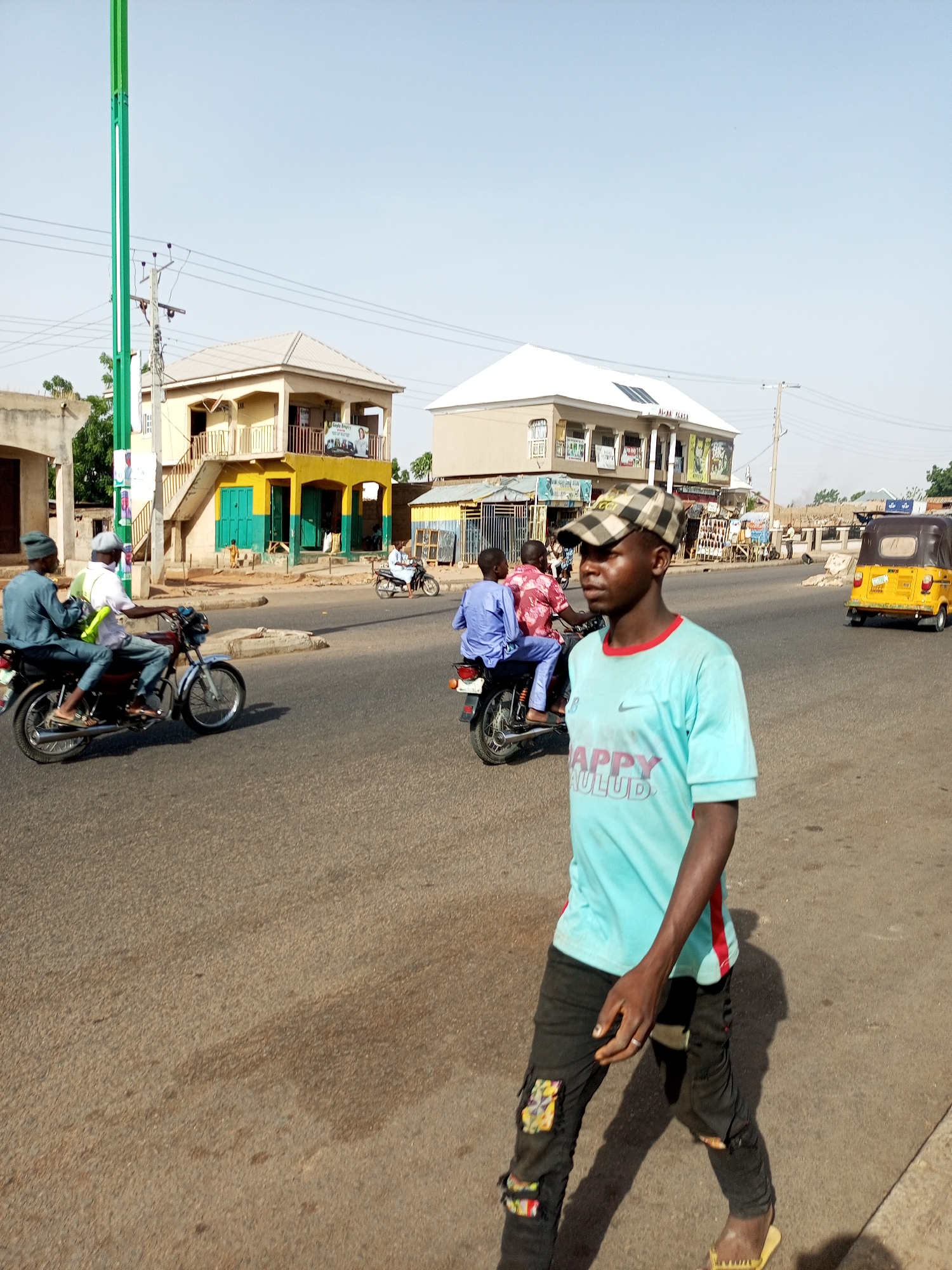 The image depicts a street scene in Gombe, Nigeria. In the foreground, a young man wearing a checkered cap, light blue t-shirt, and dark pants walks towards the camera. He has a ring on his left hand. Behind him, two motorcycles carrying passengers move along the road. A yellow auto-rickshaw (tricycle) is further down the street. Buildings line the street, one yellow with green accents and the other a beige color, each appearing to have a second story. Utility poles and wires are visible overhead. The sky is clear and pale blue. The road is asphalt.