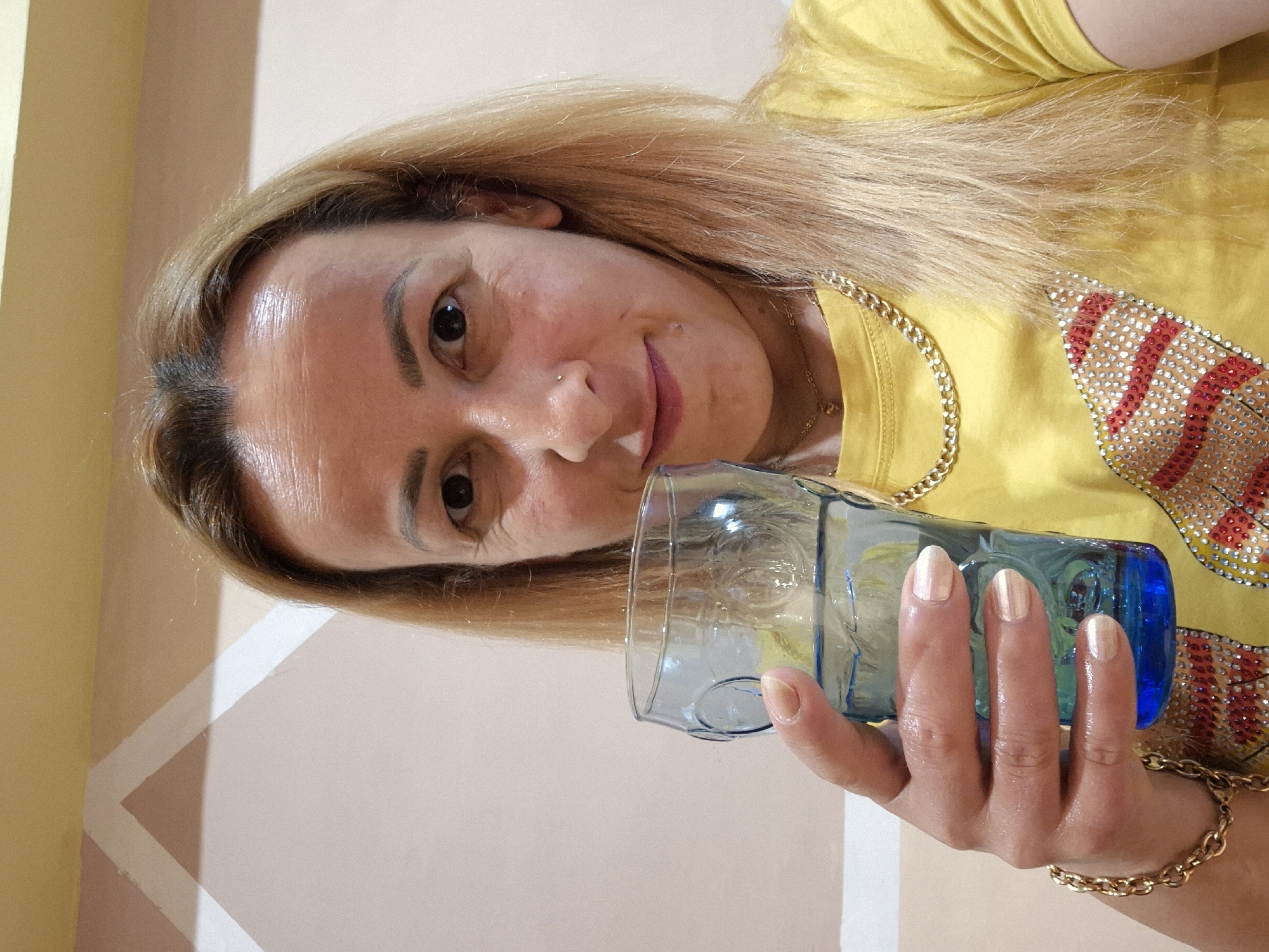A medium shot photograph captures a woman with blonde hair and fair skin, holding a blue glass of water in her right hand. She is wearing a yellow t-shirt adorned with a sparkling design of red and silver rhinestones. A gold chain necklace and a gold bracelet are also visible. The background consists of a wall with a geometric pattern in shades of beige and white. The woman is looking directly at the camera with a slight smile. The lighting appears to be indoor ambient light. The image is taken from the chest up.