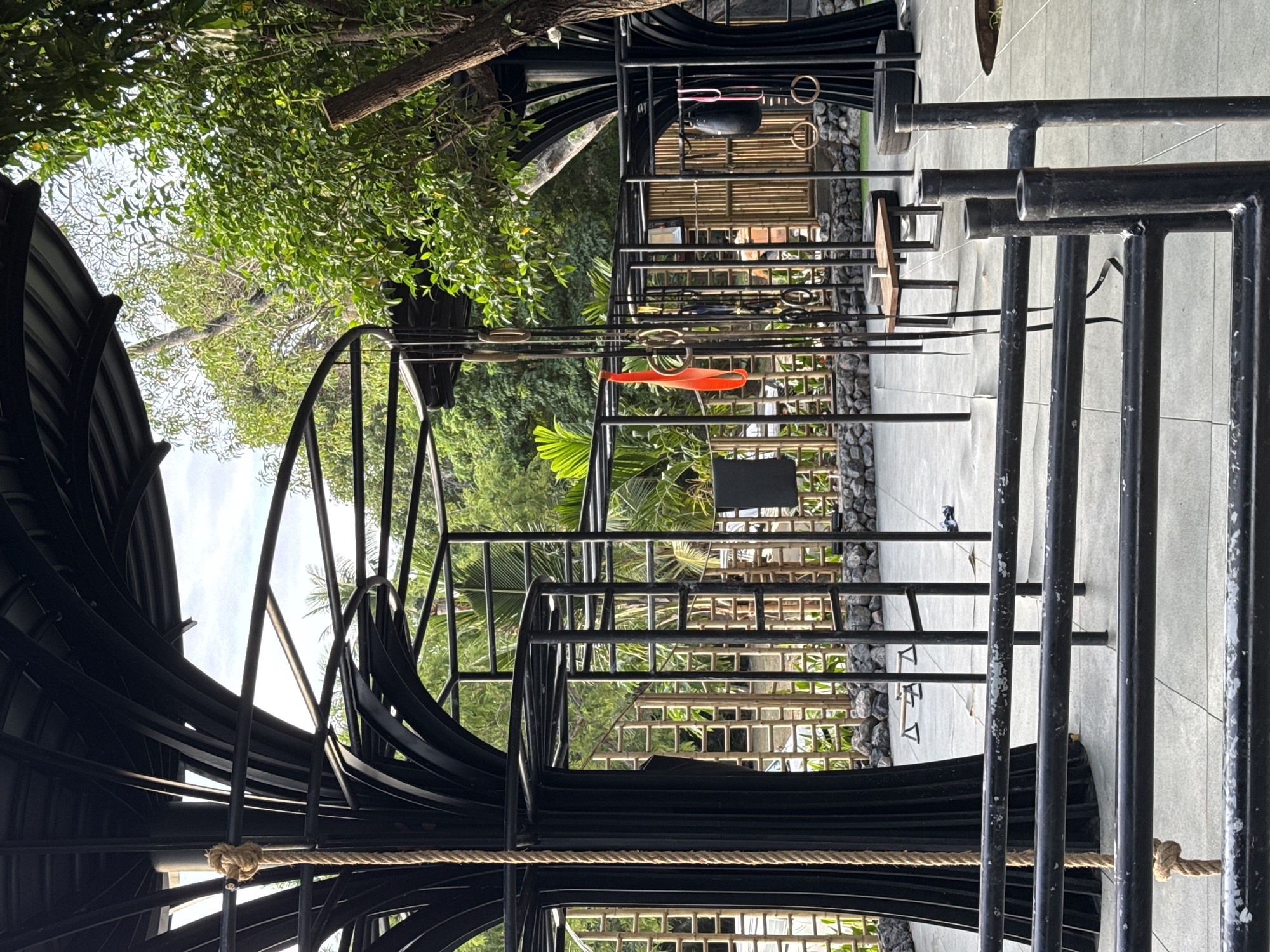 This image shows a multi-level outdoor gym facility in Baan Sri Thanu, Thailand. The structure is primarily constructed from black metal bars and features a combination of climbing frames, monkey bars, and rope elements. The ground is paved with concrete tiles. Lush greenery and trees surround the gym, blending the outdoor space with the natural environment. The perspective is from within the structure, looking outwards towards the surrounding landscape.