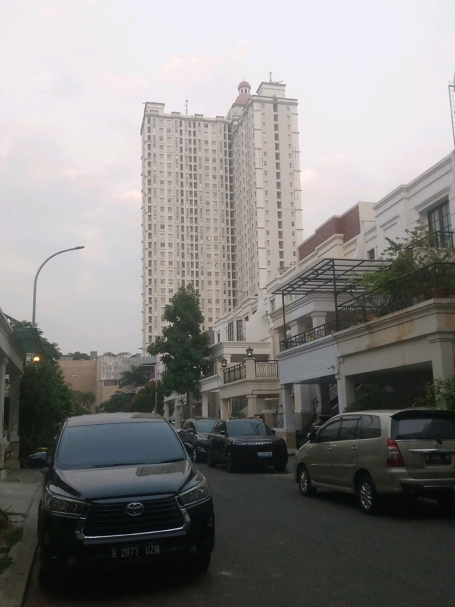 The image depicts a street scene in North Jakarta, Indonesia, dominated by a tall, modern apartment building in the background. The building is light-colored and features numerous windows arranged in a grid pattern. In front of the apartment building are several multi-story residential buildings with white facades, balconies, and dark windows. A street lamp with a curved pole is visible on the left side of the image. Several cars are parked or driving on the asphalt road. Closest to the viewer is a black SUV with a license plate starting with "B 2873 UZN." Further down the street, other vehicles including a beige minivan and a dark-colored SUV are visible. Green trees and foliage are interspersed throughout the scene, suggesting a residential or mixed-use area. The sky is overcast with a soft, diffused light, indicating it might be early morning or late afternoon. There are no people clearly visible in the image.
