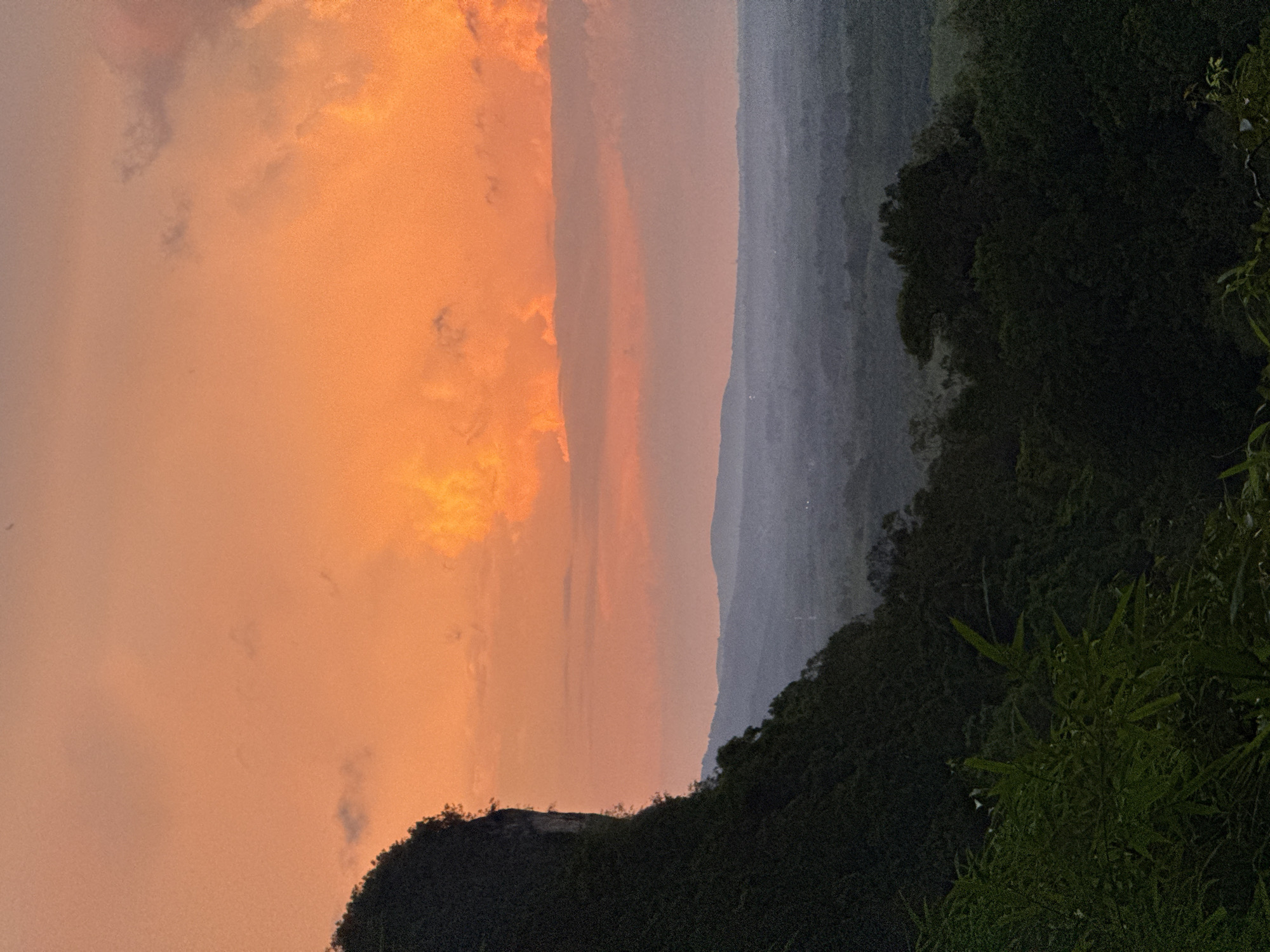 An outdoor, medium shot captures a dramatic sunset over a lush, mountainous landscape. The sky is ablaze with vibrant hues of orange and pink, with fluffy clouds scattered across the expanse. In the foreground, a steep, dark green mountainside covered in dense vegetation slopes downwards. The lower portion of the image features the shadowy canopy of a thick forest. In the distance, several layers of hazy, muted blue mountains recede into the horizon. The overall scene suggests a remote, natural setting, potentially a viewpoint overlooking a valley. The specific location is identified as Ban Khao Tang, Thailand. There are no people or discernible man-made structures within the frame.
