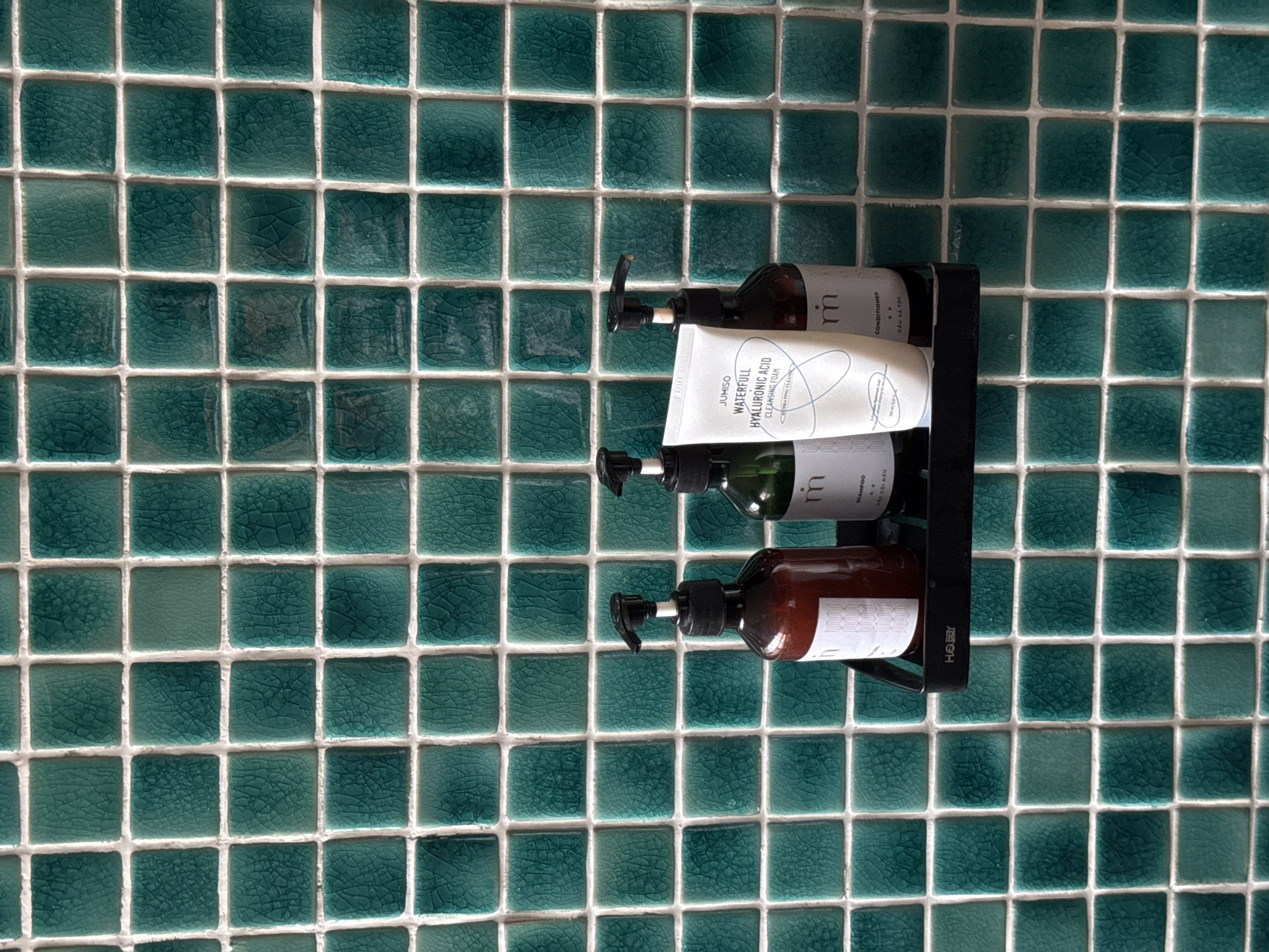 The image shows a black shelf attached to a wall covered in square, teal-colored tiles. On the shelf, from left to right, are three bottles and one tube of product. The first bottle is a dark brown glass bottle with a black pump dispenser. It has a white label with a pattern of vertical wavy lines. The second bottle is green glass with a black pump dispenser and a white label. The third bottle is also dark brown glass with a black pump dispenser and a white label. In front of the green bottle and to the right of the dark brown bottle is a white tube with a black cap. The tube has text that reads "JUMISO WATERFULL HYALURONIC ACID CLEANSING FOAM". The shelf has the word "HOBBY" printed on its front in white. The image is taken from a slightly elevated angle, looking down at the shelf and its contents against the tiled background. The location is South Quy Nhon Ward, Vietnam.