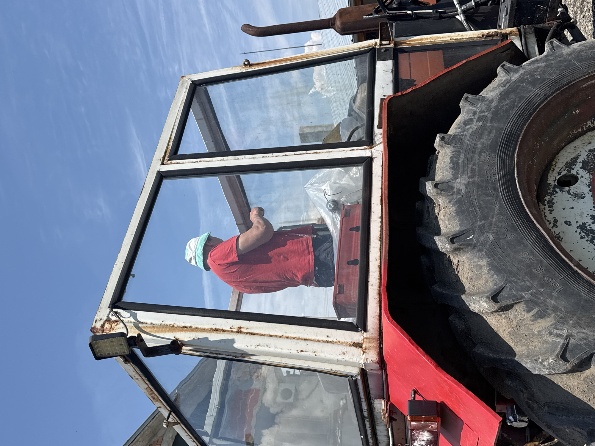 The image depicts an outdoor scene in Sarnevo, Bulgaria, featuring a weathered red tractor with a white and rusty cab. A person, viewed from behind, is situated inside the cab.
The individual wears a red short-sleeved t-shirt and a light blue and white bucket hat. Their hands are engaged with an unseen object or structure inside the cab, suggesting an action of maintenance or adjustment. Visible within the cab are a red toolbox and items covered by a white plastic sheet or bag.
The tractor is an older model, characterized by significant wear and rust on its white metal cab frame, particularly at the joints and where an aftermarket LED work light is mounted on the front left corner of the roof. The cab's glass windows reflect a bright blue sky with sparse, wispy clouds, as well as portions of distant buildings and an air conditioning unit. The prominent rear tire, marked "155 R 38" and "Radial EPR," is large, heavily treaded, dirty, and shows wear. Its white rim is chipped and rusted. A rusty, upward-curving exhaust pipe is visible on the right side of the tractor. The red metal fender over the rear wheel is also worn. The scene is illuminated by strong daylight under a clear sky.