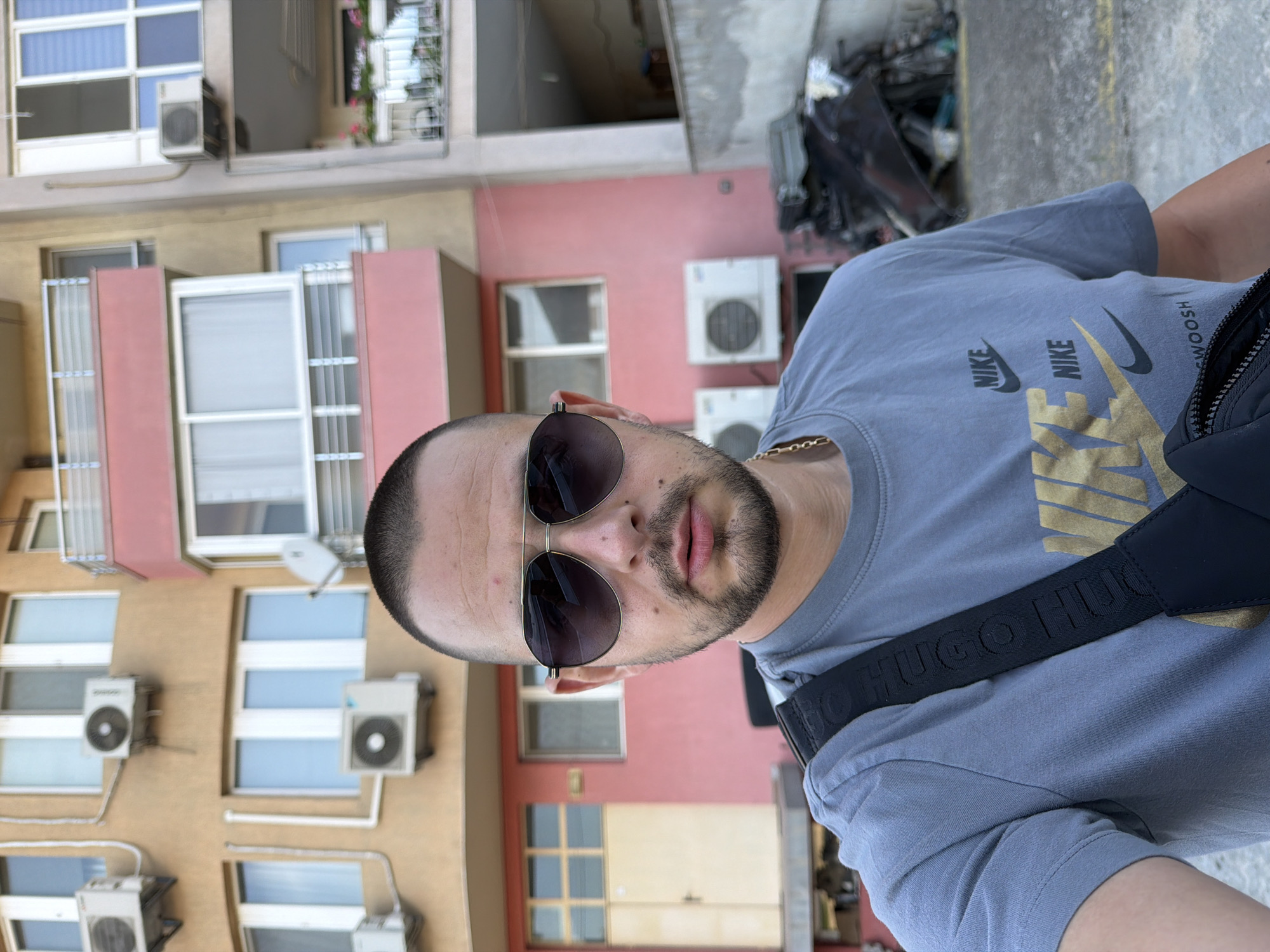 A medium shot shows a man facing the camera, wearing sunglasses and a gray Nike t-shirt with a black strap bag across his chest. Behind him, a multi-story building with beige and pink facade features several air conditioning units and windows. The building appears to be located in Burgas, Bulgaria, based on the visual style of the architecture and the presence of air conditioning units commonly seen in the region. The ground appears to be a concrete or asphalt surface, with some debris and what looks like discarded materials in the background. The lighting suggests it is daytime, with natural light casting shadows. The man's expression is neutral, and he is looking directly into the camera.