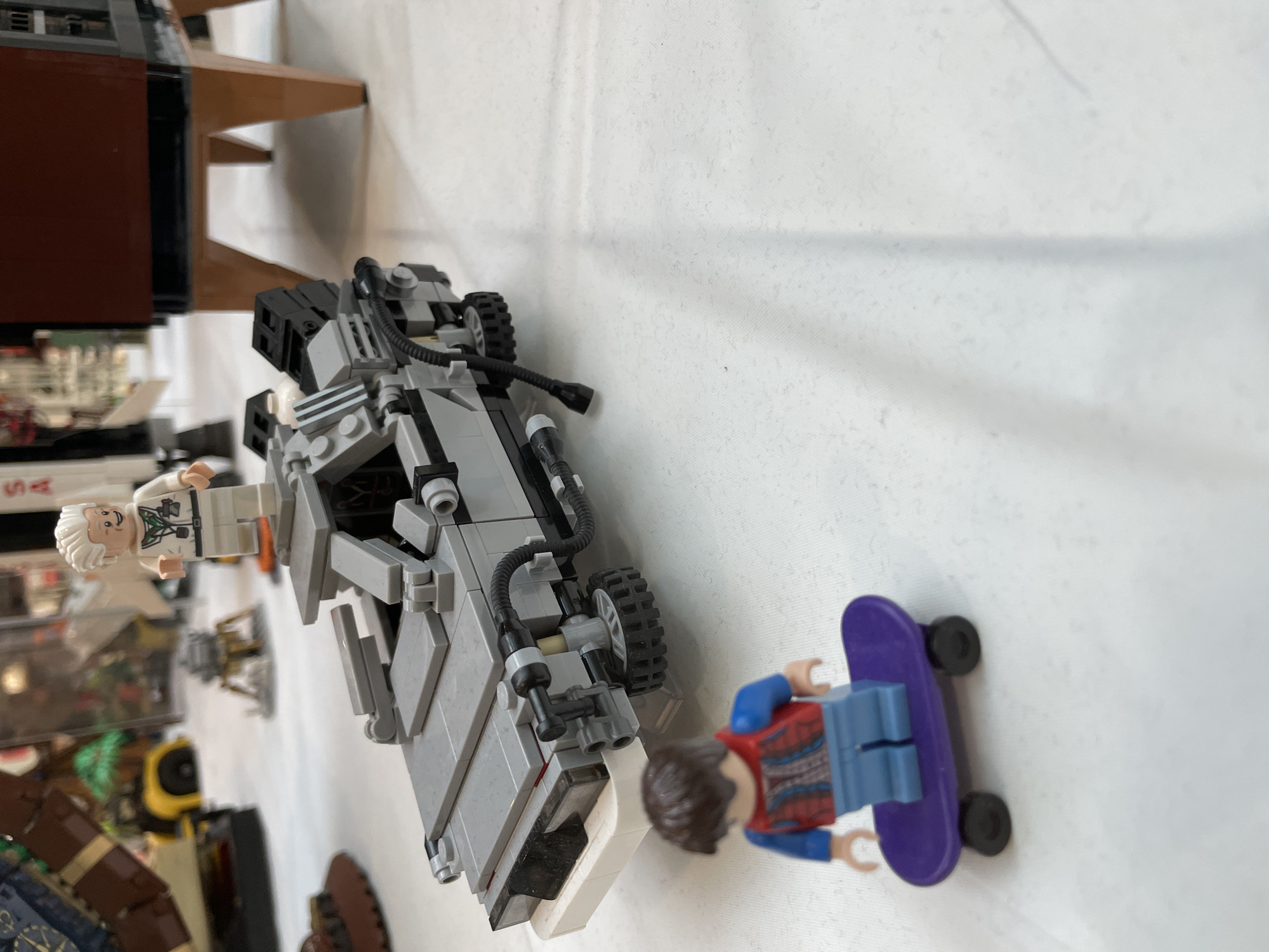 The image shows a close-up view of Lego figurines and models on a white, crinkled surface, resembling a tablecloth. In the foreground, a Lego figurine of Marty McFly from "Back to the Future" is depicted riding a purple skateboard. Behind him is a Lego model of the DeLorean time machine from the same movie franchise. To the right of the DeLorean, a Lego figurine resembling Doctor Emmett Brown stands on a grey platform. In the background, other Lego models and figurines are visible, including what appears to be a rocket and a yellow vehicle. The lighting suggests an indoor setting with diffused light, possibly from a window or overhead lamps. The overall scene evokes a display of Lego creations, likely at an exhibition or event. There is no indication of the specific location being in Sofia, Bulgaria.