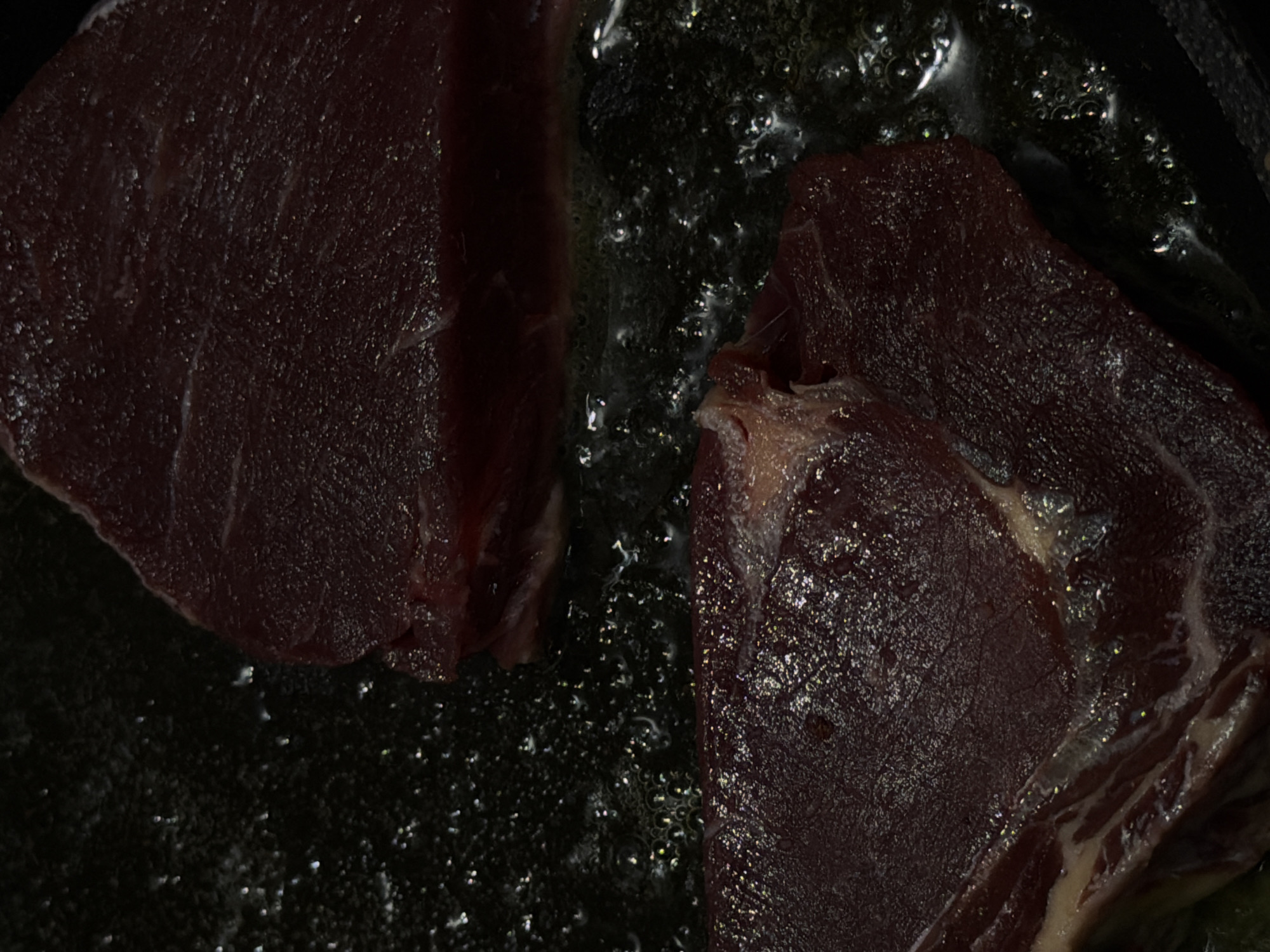 Two pieces of raw, dark red meat are being cooked in a pan filled with bubbling liquid. The meat appears to be browning slightly. The image does not provide any information about North Kuta, Indonesia, people, interactions, or the environment outside the pan.