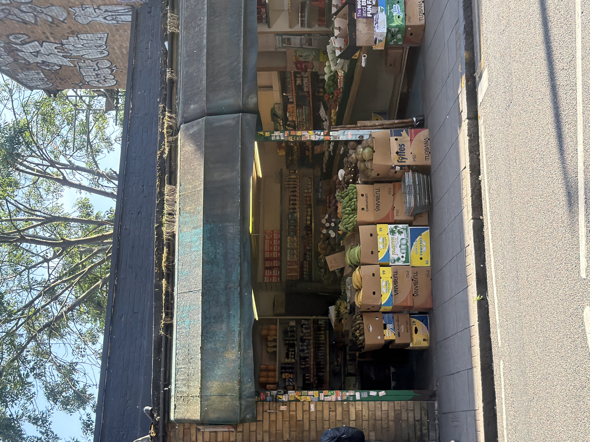 The image captures a street-level view of a small corner shop with a dark awning, located in London, United Kingdom. The shop, constructed from brown brick, features a display of fresh produce, including bananas, coconuts, and other fruits and vegetables, arranged in cardboard boxes and crates outside its entrance. A portion of the shop's interior, stocked with shelves of various products, is visible through the open doorway. In the background, a building adorned with graffiti and scaffolding suggests ongoing construction or renovation. The pavement in front of the shop is paved with concrete slabs, and the street beyond is asphalt. A person, partially visible on the far left of the frame, is wearing a blue jacket. The overall scene depicts a typical urban commercial environment with a focus on a local grocery store.