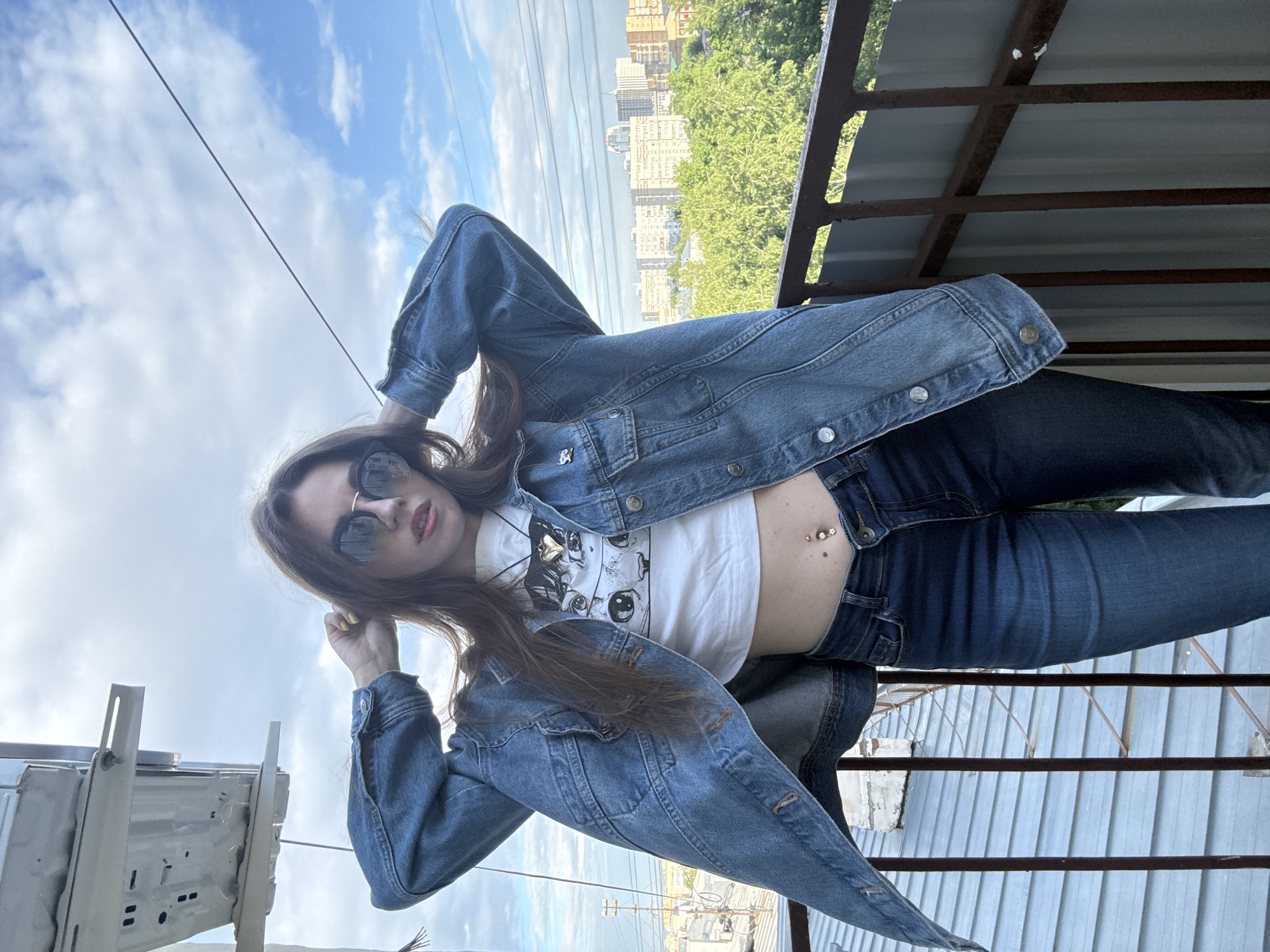 A young adult female with brown hair and sunglasses is reclining on a metal structure. She is wearing a white crop top with a cartoon image, a blue denim jacket, and blue jeans. Her navel is pierced. The background features a city skyline with buildings and trees, under a cloudy sky. The location is Moscow, Russia.