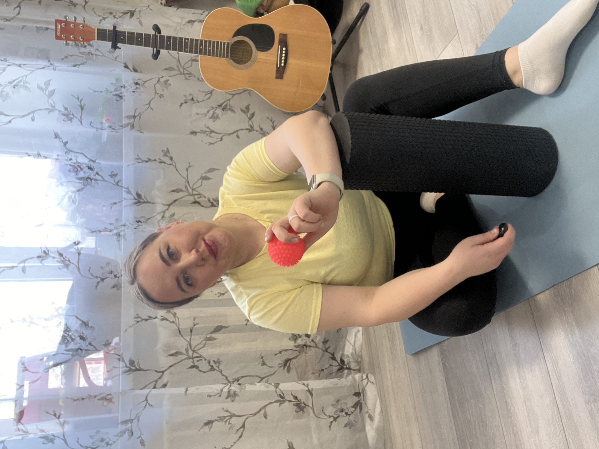 This image, taken indoors in Chaikovsky, Russia, features a woman seated on a light blue yoga mat on a light wooden floor. She is looking directly at the camera with a neutral to slightly smiling expression. The woman has light-colored hair and is wearing a yellow short-sleeved t-shirt, black leggings, and white socks.
On her left wrist, she wears a light-colored smartwatch. In her right hand, she holds a small, bright red, spiky massage ball. A large, black, textured foam roller is positioned vertically next to her legs on the mat, suggesting an activity related to fitness, stretching, or self-massage. A smaller, dark, round object is also visible near her left hand.
The background shows sheer white curtains with a repeating pattern of grey and black floral branches, indicating a window behind them. To the left side of the frame, an acoustic guitar with a light wood finish is propped upright on a stand. The lighting in the room appears bright and natural, suggesting it is daytime. There is no visible text in the image. The scene portrays a casual moment, possibly before or after a personal wellness routine.