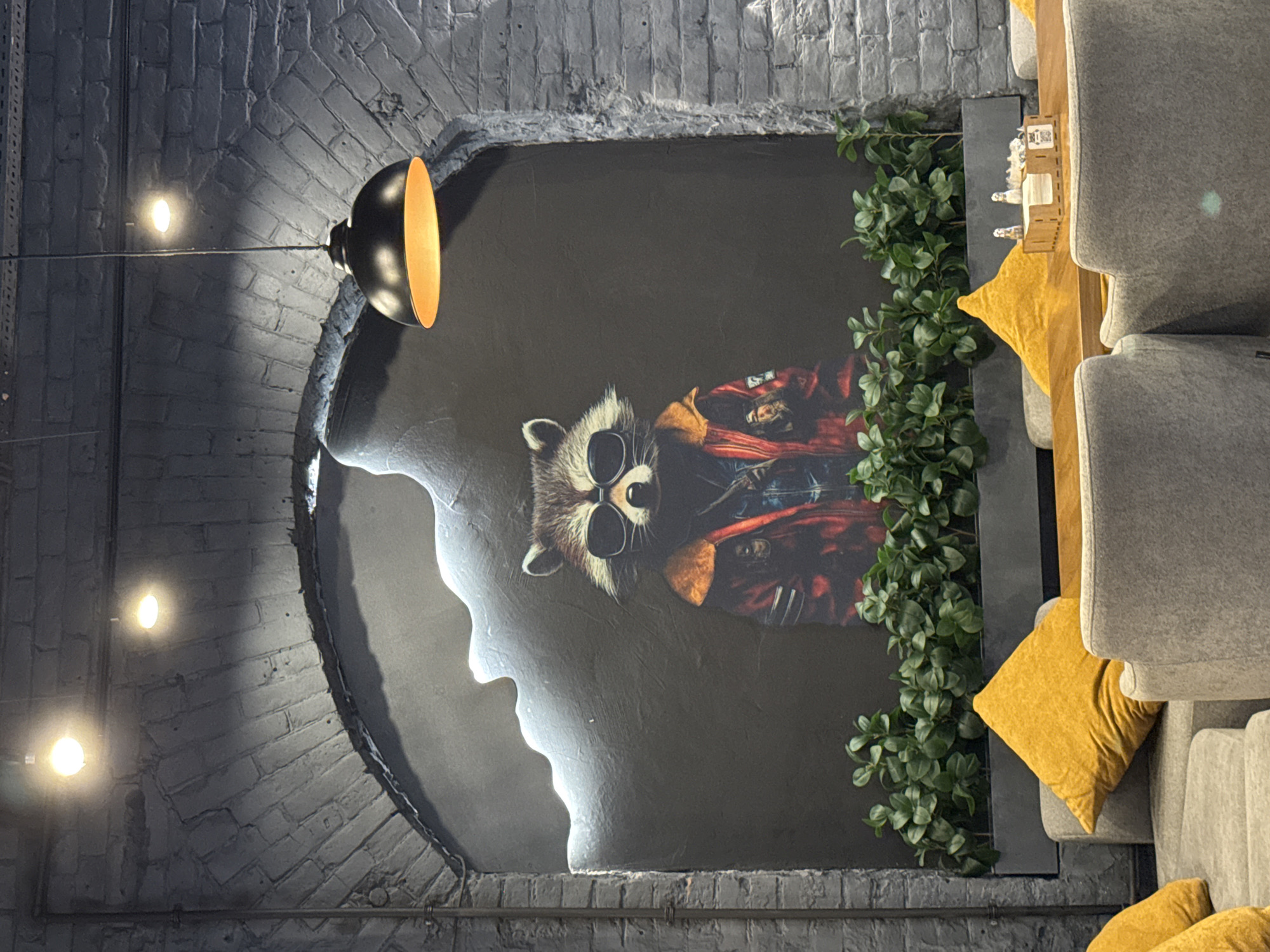 The image shows an interior space, likely a cafe or lounge, in Samara, Russia. A large, stylized mural of a raccoon wearing sunglasses and a red and blue jacket is the central focus. The raccoon appears to be cool and relaxed, possibly smoking or holding something in its paw.
The mural is set within an arched alcove against a dark gray background. The alcove's edge is highlighted with a bright white light, creating a dramatic contrast. To the right of the mural, lush green artificial plants cascade down, partially obscuring the artwork and adding a touch of nature to the artificial setting.
The surrounding walls are made of dark gray brick, contributing to the dimly lit, industrial-chic aesthetic. Several pendant lights are visible: one large, metallic dome-shaped light with a warm orange interior hangs from the upper left, casting a strong downward glow. Two smaller, circular lights are also visible on the brick wall to the left, emitting a soft, yellowish light.
In the foreground, to the right of the mural and plants, are plush gray seating areas, likely sofas or armchairs. Bright yellow cushions are placed on these seats, adding pops of color and warmth to the scene. A small wooden shelf unit sits on a surface, holding a few decorative items, including what appear to be small figures and possibly some type of business cards or signage. The overall ambiance suggests a modern, trendy, and inviting space designed for relaxation and socializing. The lighting indicates it's likely evening or the interior lights are the primary source of illumination.