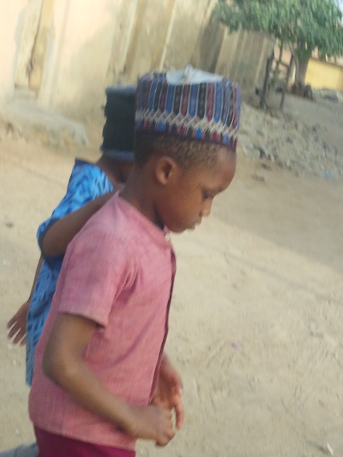 Two young individuals are captured outdoors on a light-colored sandy or dirt surface in Garun Gawa, Nigeria. In the foreground, a child with dark skin, wearing a pink short-sleeved collared shirt and a multi-patterned cap (kufi) with blue, red, black, and white elements, stands with their head bowed, looking downwards. Behind this child, partially obscured, is another individual wearing a blue patterned garment and a dark head covering. A light-colored, possibly concrete or mud-brick, building structure is visible in the left background, while a tree with green foliage is present in the upper right background. The scene is illuminated by natural light.