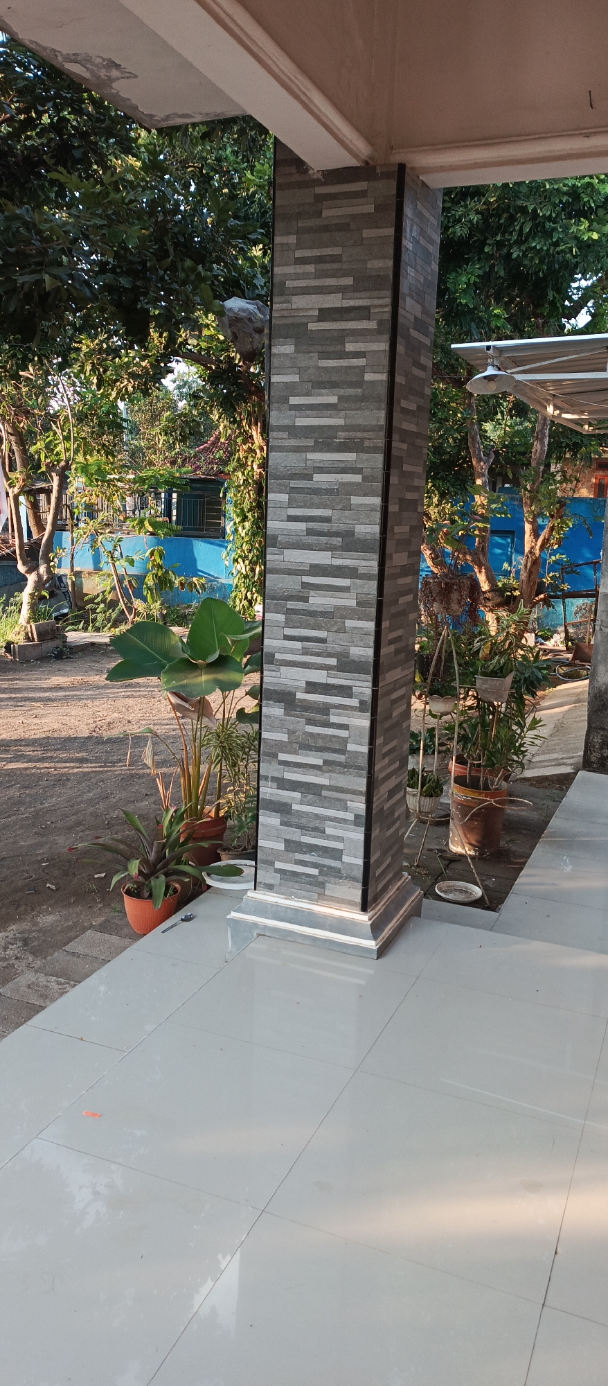 The image depicts an outdoor scene at a residence in Jatisari, Indonesia. A tiled pillar, clad in a pattern of grey and white rectangular tiles, stands prominently in the foreground. To the left of the pillar, several potted plants are arranged on a light-colored tiled patio. These plants include a large-leafed tropical plant and a smaller, spiky plant in an orange pot. Beyond the patio, a dirt pathway is visible, leading towards a bright blue wall of a building. Lush green trees with dense foliage surround the area, suggesting a tropical environment. Sunlight casts shadows on the ground, indicating it is likely daytime. The overall impression is of a well-maintained outdoor space adjacent to a dwelling.