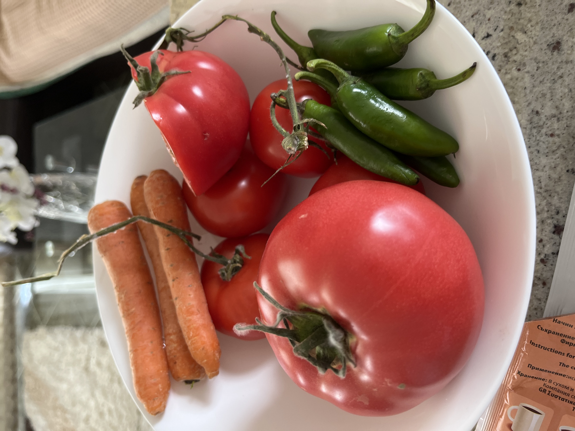 A top-down shot shows a white bowl filled with fresh produce: ripe red tomatoes (one partially sliced), small orange carrots with stems still attached, and a cluster of green chili peppers. A packet with text in Cyrillic and English is partially visible to the side of the bowl, suggesting the image was taken somewhere where both languages are used. The background consists of a glass tabletop and other blurry objects, including possibly a white sofa or cushion. There is nothing indicating that this scene is located in Zlatograd, Bulgaria.