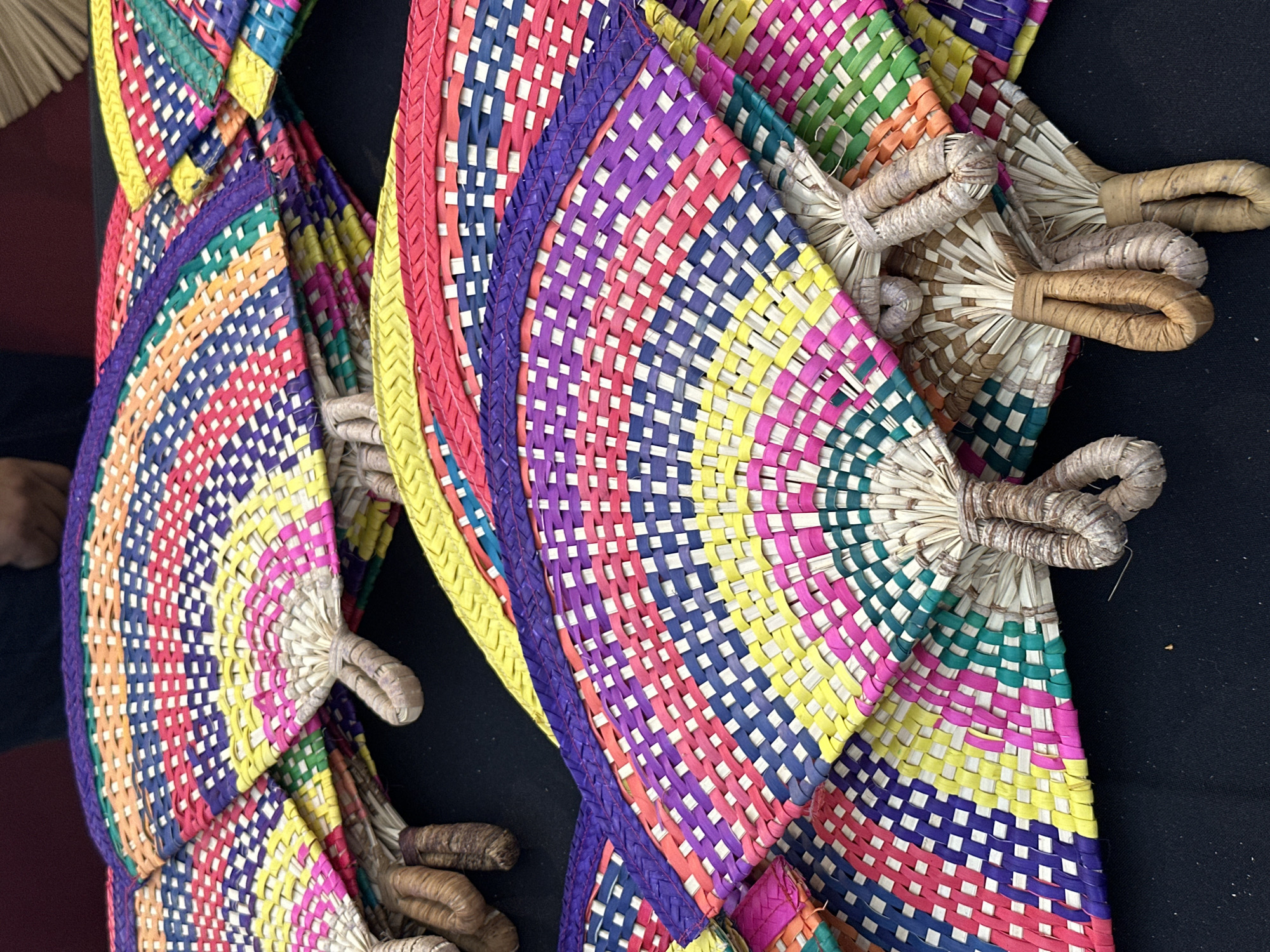 The image, captured in Mexico City, Mexico, presents a close-up, eye-level view of numerous handcrafted, vibrantly colored woven palm leaf fans displayed on a dark, likely black, textile surface. Approximately a dozen fans are visible, stacked and overlapping, showcasing their distinctive semicircular, fanned-out shapes. Each fan is constructed from tightly woven, dyed natural fibers, featuring intricate patterns composed of segmented stripes and checkerboard motifs in a broad spectrum of colors including red, pink, yellow, various shades of green, blue, purple, orange, and white. The fan handles are crafted from undyed, light brown, braided, or wrapped natural fibers, forming a loop at the base. In the blurred upper background, sections of dark clothing and a human hand are partially visible, indicating the presence of individuals in proximity to the display, consistent with a market or craft fair setting.