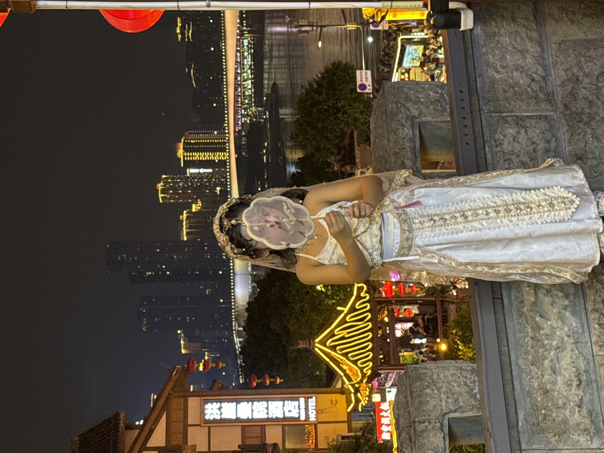 A young woman wearing a traditional Chinese costume and a veil stands on a stone wall at night. She holds a fan in front of her face. In the background, a river reflects the lights of a city skyline with numerous tall buildings and a bridge. The scene appears to be in Yuzhong District, China, a location known for its vibrant night life and architecture. Buildings with traditional Chinese architectural elements are visible to the left, along with illuminated signs and red lanterns.