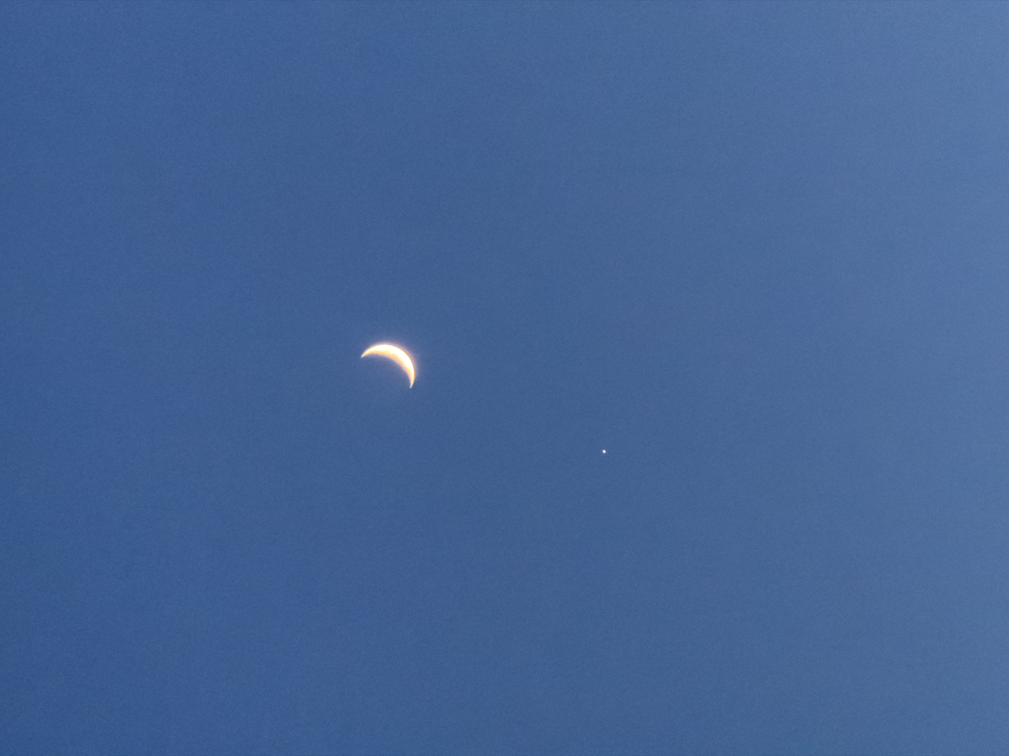 A daytime photograph depicts a thin crescent moon accompanied by a single, bright star-like celestial body against a clear, blue sky. The moon is positioned on the left side of the frame, exhibiting a slender arc of reflected sunlight. To the right and slightly below the moon is a single point of light, possibly a planet or bright star. The scene is absent of any earthly features or indications of the Krasnoyarsk location.