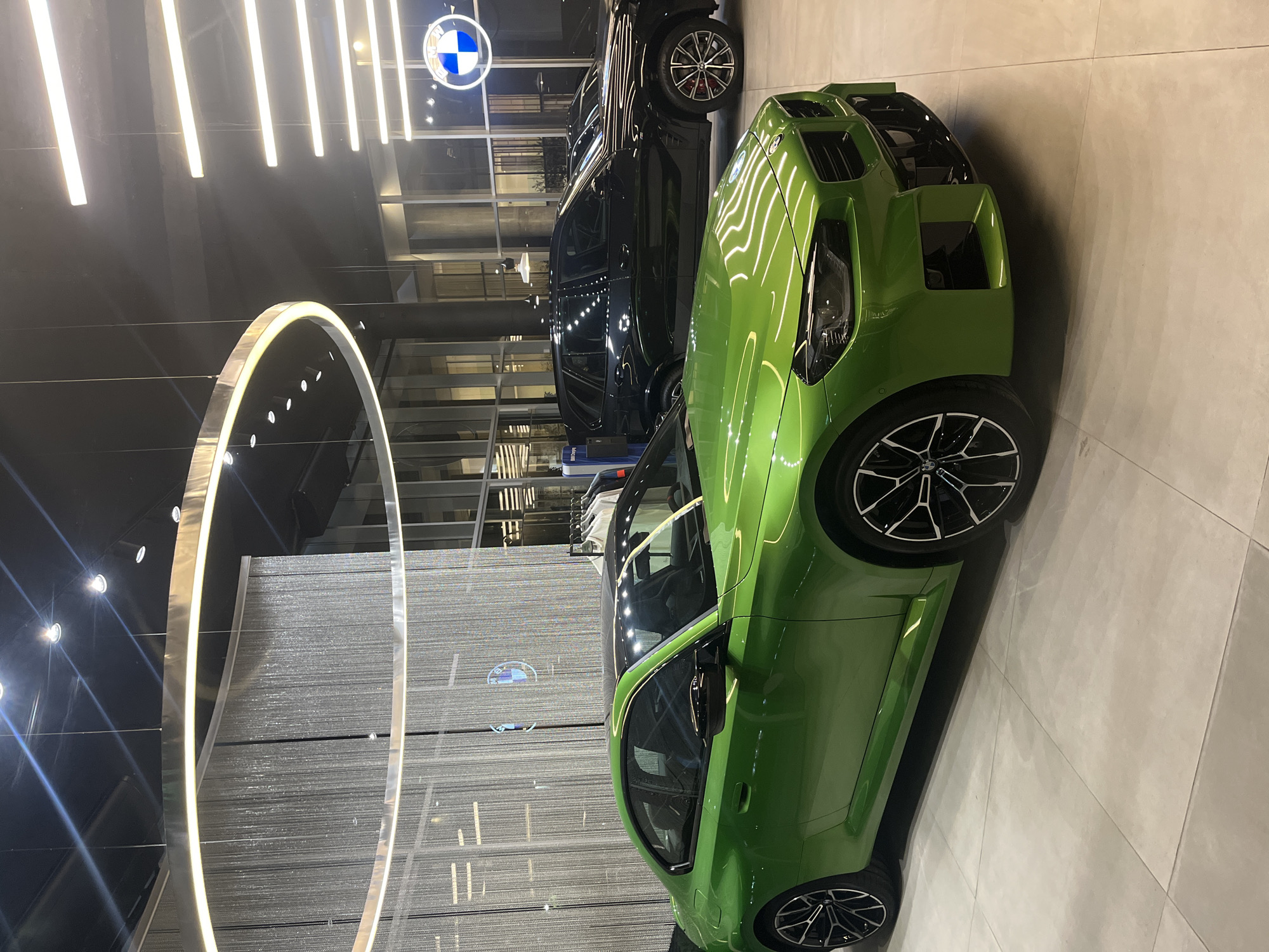 The image displays a modern BMW car showroom, likely located in Buenos Aires, Argentina, as indicated by the context. The scene is dominated by two high-end BMW vehicles. In the foreground and positioned to the right, a vibrant bright green BMW sports car, possibly an M3 or M4 model, is prominently featured. Its distinct front fascia, sleek side profile, and multi-spoke black and silver alloy wheels are clearly visible, with ample light reflecting off its glossy surface. Behind it, further to the left, is a black BMW SUV, appearing to be an X5 or X7, also equipped with characteristic alloy wheels and red brake calipers.
The showroom's interior is brightly lit and presents a contemporary, upscale design. The floor consists of large, light-colored square tiles. Overhead, numerous linear LED light strips illuminate the space, creating bright reflections on the cars and glass surfaces. A distinctive architectural element is a large, illuminated circular ring structure situated in the mid-ground. In the background, large glass panels offer glimpses of other parts of the showroom, while a textured wall provides depth. The iconic blue and white circular BMW logo is visible in the upper left background, confirming the brand. No people are present, emphasizing the vehicles and the clean, sophisticated environment. The strong artificial lighting suggests an indoor setting, possibly during evening hours.