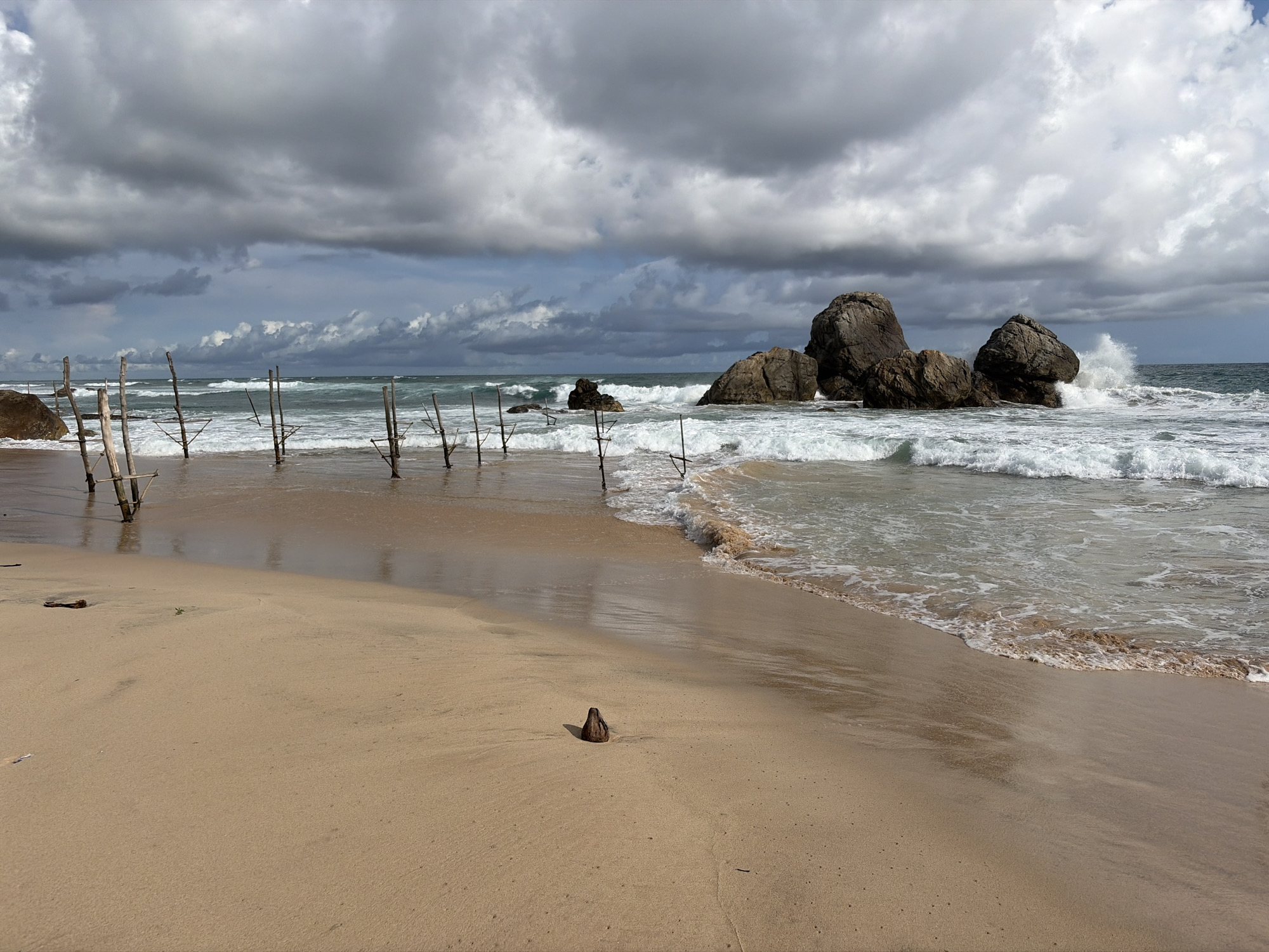 The image shows a beach in Habaraduwa, Sri Lanka, on a cloudy day. In the foreground, there's a sandy beach with a small piece of driftwood. Further into the scene, the beach meets the ocean. Sticks stand in the shallow water near the shore. Offshore, several large rock formations are visible, with waves crashing against them. The sky is filled with dark, heavy clouds. No people are visible in the frame.
