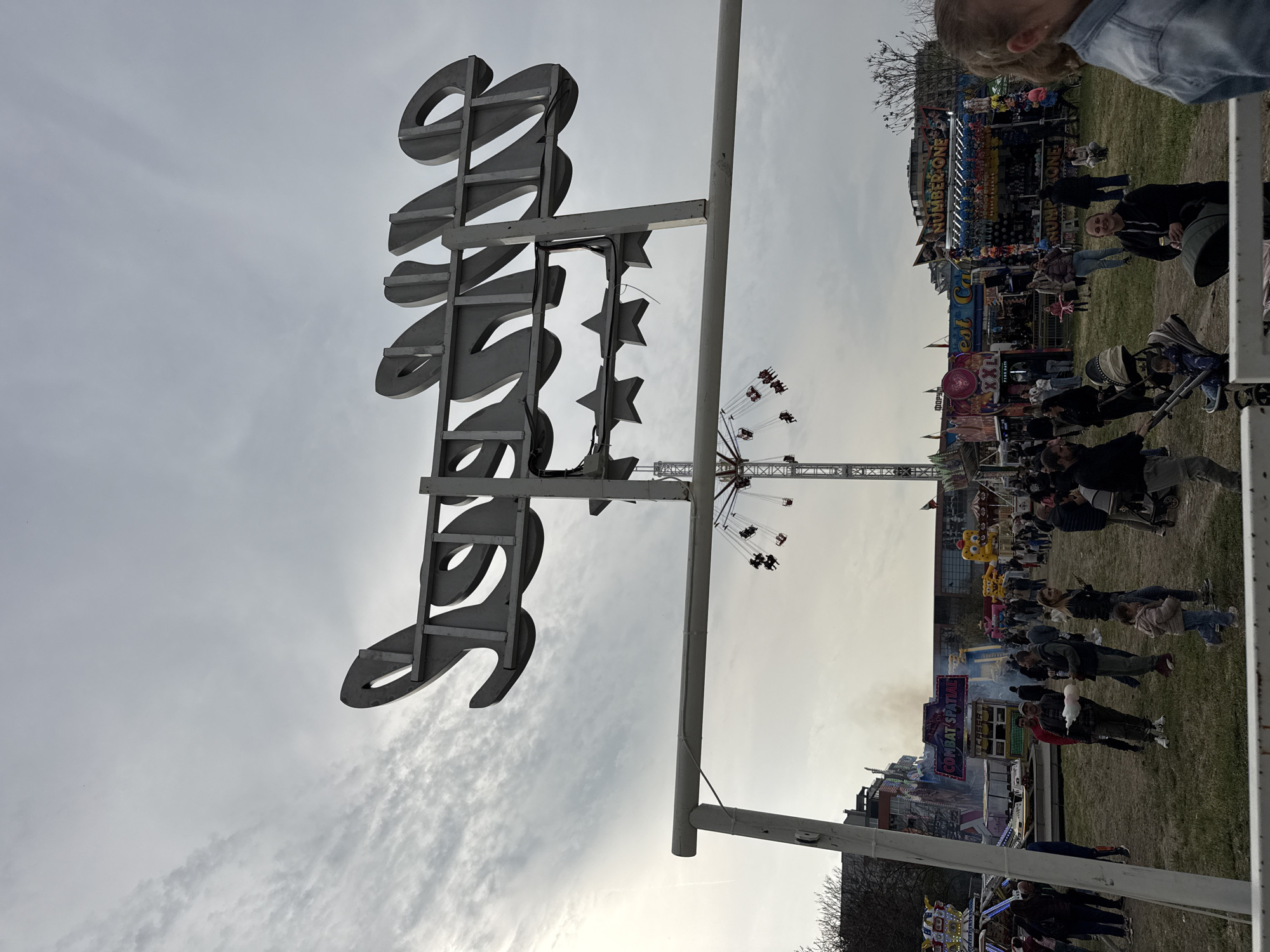 The image depicts a fair or carnival in Plovdiv, Bulgaria. The fair includes rides such as a swing ride. A sign with stars is in view. People, including adults and children, are walking around the fair. Game booths are set up. The sky is overcast.