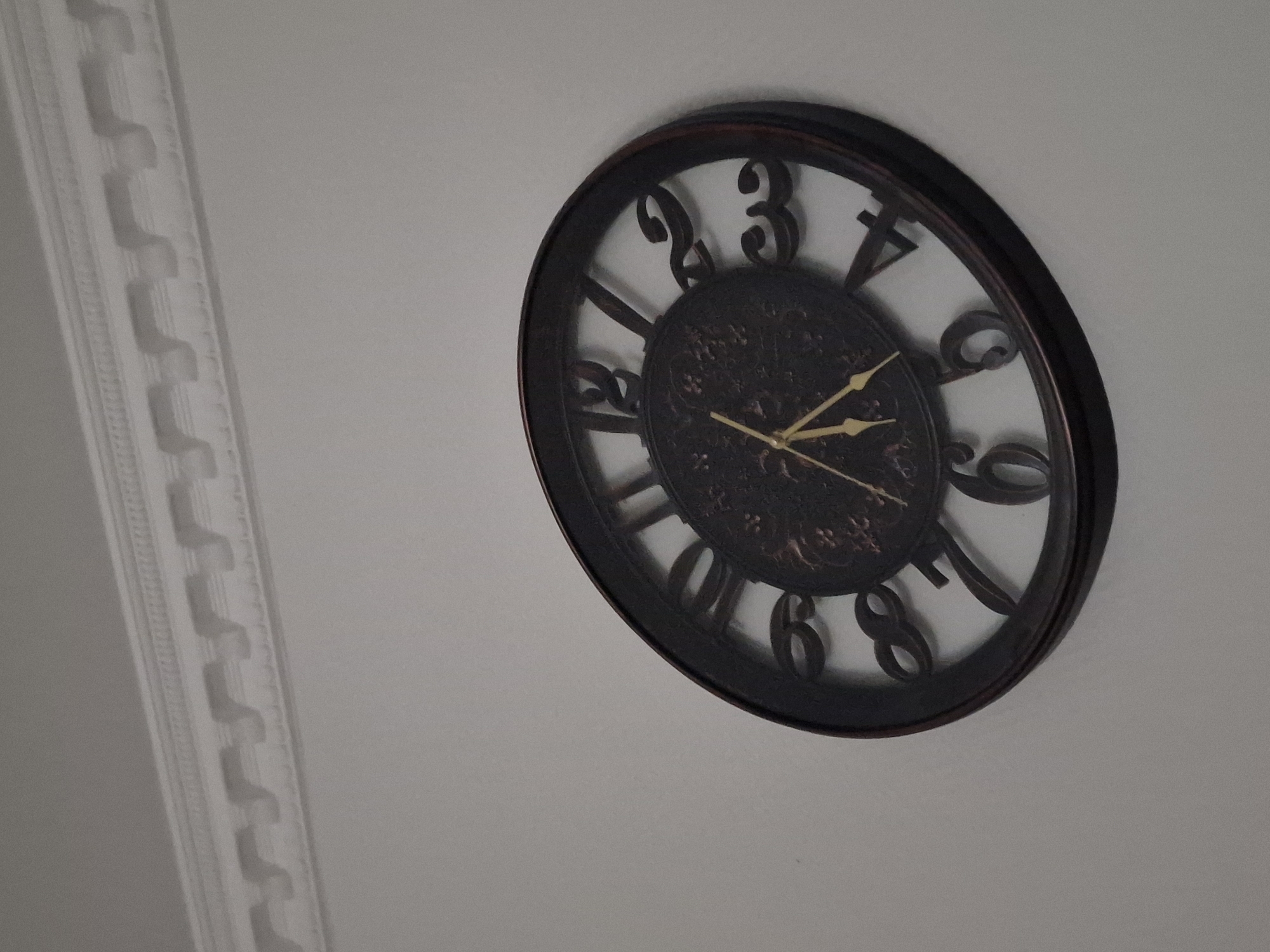 This media file contains a photograph of a dark-framed, analog wall clock with Roman numerals. The clock face has an ornate, dark design. The hour hand points to approximately the 2, and the minute hand points to approximately the 4. The clock is mounted on a light-colored wall. To the left of the clock, a decorative white molding runs vertically down the wall. The molding has a repeating geometric pattern. The overall setting appears to be indoors, likely a room in a home or office. There is no visible text in the image. The lighting suggests it is daytime, and the scene is static with no activity taking place.