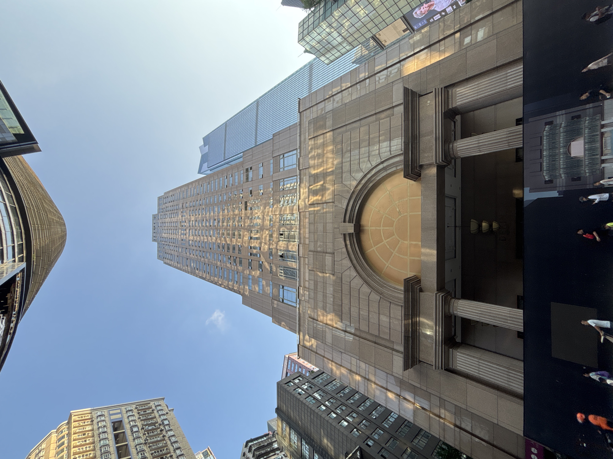 The image is a low-angle shot of a modern cityscape, dominated by towering buildings and a clear blue sky. In the foreground, a large, light-brown building with a stone facade features a grand entrance with two fluted columns and an arched window above. The facade is decorated with geometric patterns and reflects sunlight, creating diagonal highlights. Behind this building, a taller, slender skyscraper with a reflective blue glass exterior rises into the sky. To the left, a dark-colored building with a grid of windows is visible. In the background, other buildings of various heights and architectural styles can be seen. At the very bottom of the image, a black barrier or wall runs horizontally, with several people standing or walking in front of it. One person in an orange top is clearly visible on the far left, and another person in a white shirt is pushing a cart. A banner with a portrait of a person is attached to the building on the right. The scene appears to be in a bustling urban environment, possibly Yuzhong District, China, given the modern architectural style and density of skyscrapers. The lighting suggests it is daytime, with the sun casting long shadows.