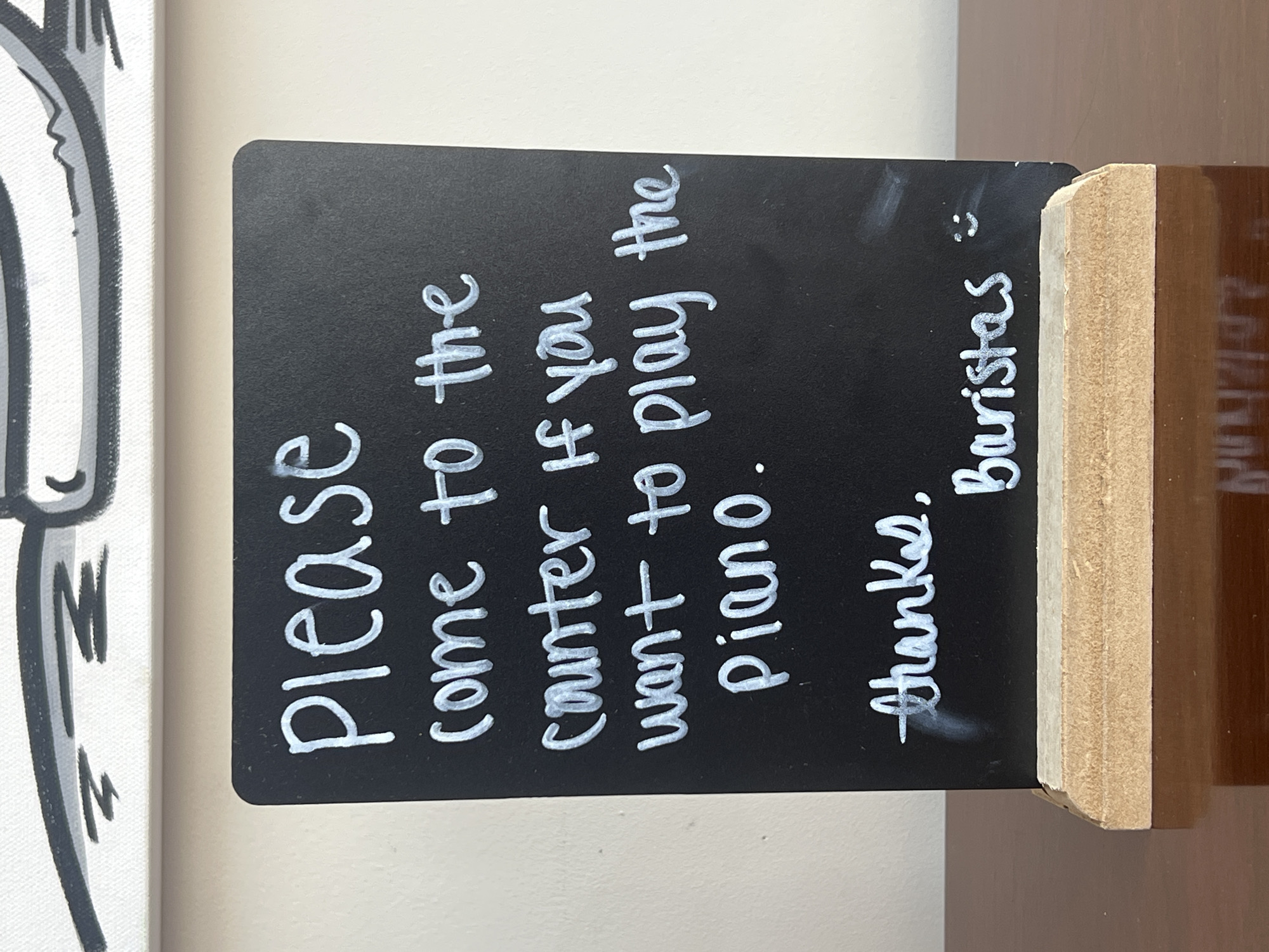Here is a detailed description of the image:
The image shows a small, chalkboard-style sign with white lettering. The sign reads: "Please come to the counter if you want to play the Piano. Thanks. Baristas :)" The sign is propped up in a small, wooden stand. To the left of the sign is a portion of a white canvas with a black and white illustration. The background appears to be a plain wall or countertop.
Based on the sign's content and the "Baristas" signature, the scene is most likely located in a coffee shop or cafe. No people are visible in the image. Given the request for location information, the assumption is that this photograph was taken within the City of Newburgh, United States of America, but no specific landmarks or features in the image confirm this.