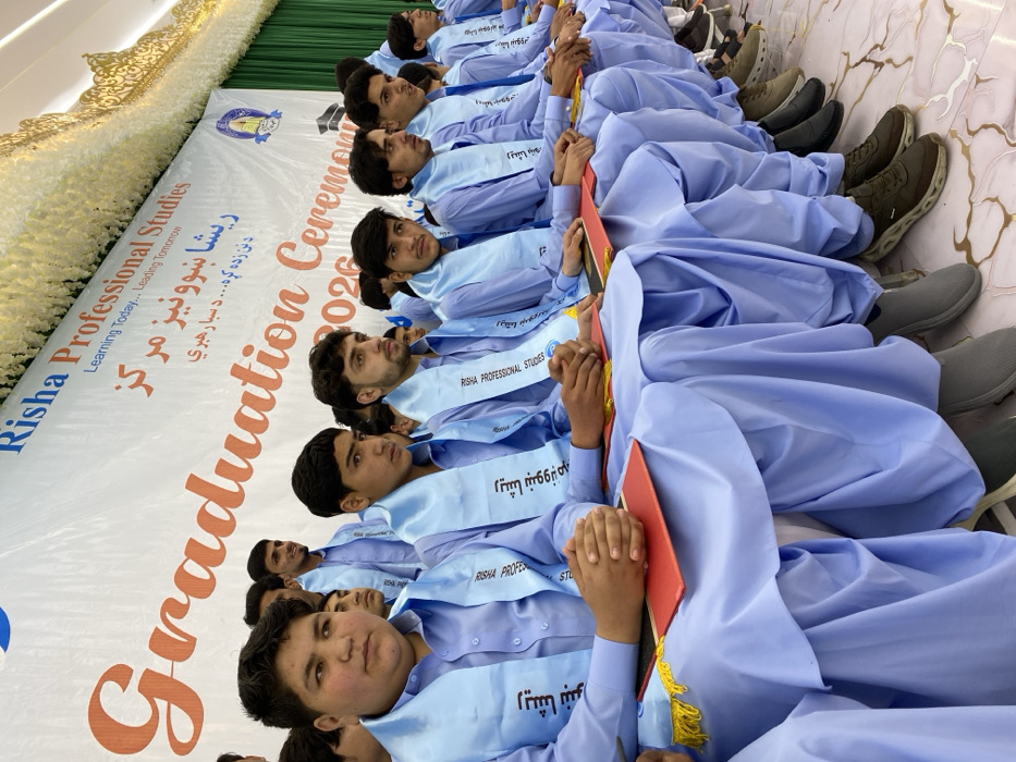 A group of young men are seated in rows at a graduation ceremony for Risha Professional Studies. They are wearing light blue robes and stoles with text written in both English and what appears to be a local dialect. Each student is holding a red diploma or certificate. A banner behind them displays the name of the institution, "Risha Professional Studies," along with the phrases "Learning Today... Leading Tomorrow" and "Graduation Ceremony 2026." The text is also written in the local dialect.