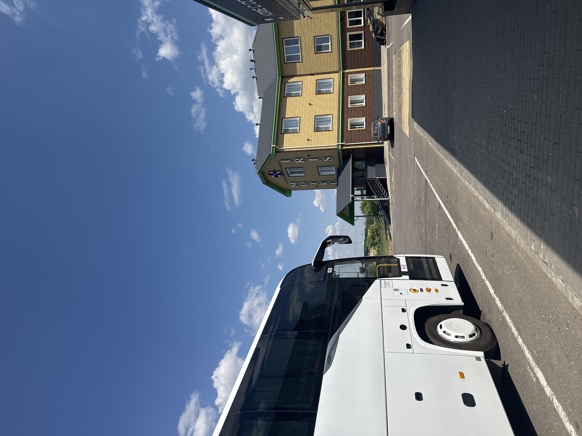 The scene depicts a white bus parked in a paved parking area in Bol'shie Nyrsi, Russia. The bus is positioned in front of a three-story building, the IRIS Hotel Motel. The building has a combination of yellow and brown wooden siding, with white-framed windows. A car is also parked near the hotel entrance. The sky above is blue with scattered clouds.