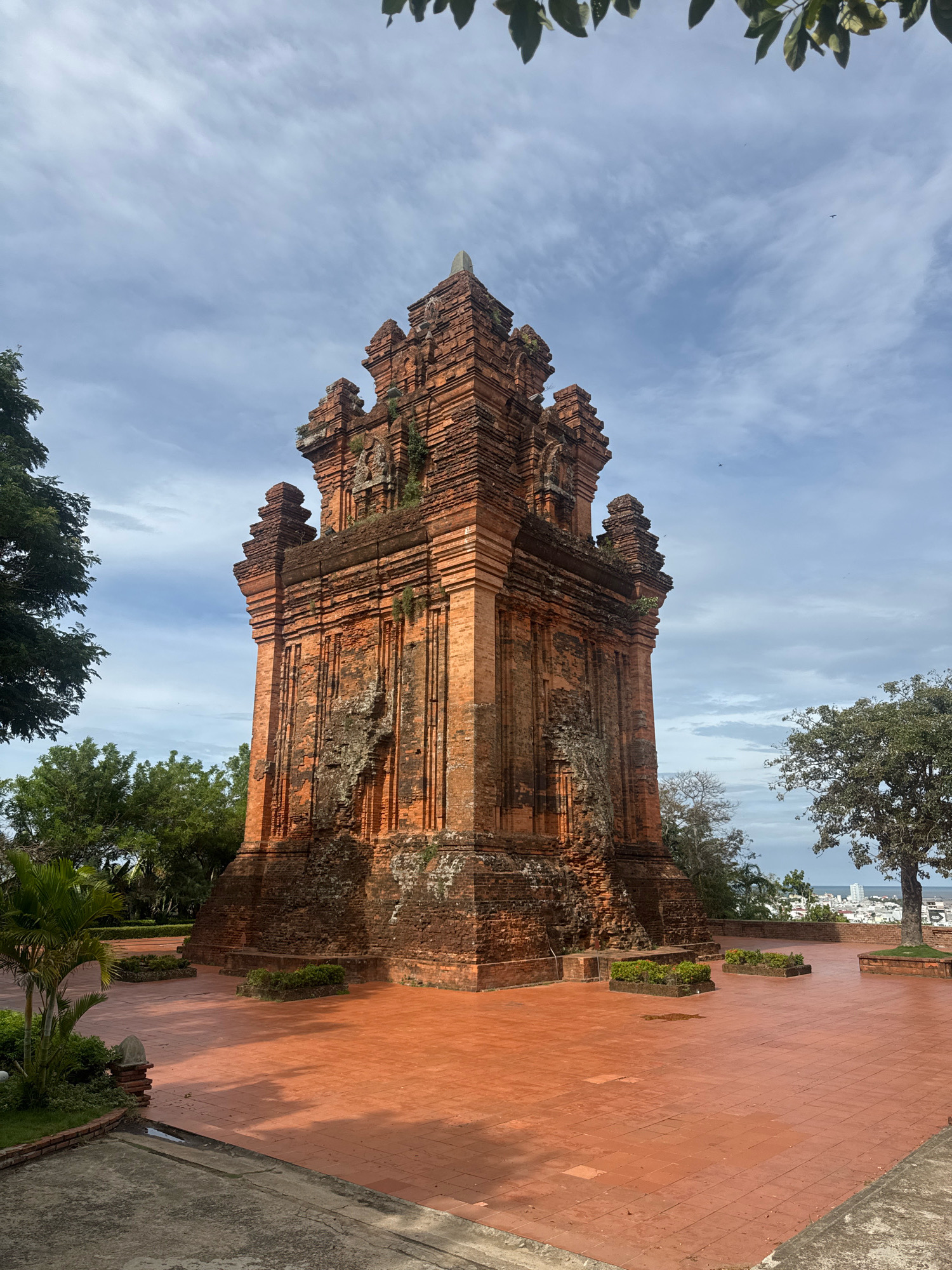 The image depicts the Po Nagar Cham Towers in Tuy Hoa, Vietnam. These ancient brick towers, constructed in the Champa architectural style, stand tall against a cloudy sky. The foreground features a vast expanse of reddish-orange tiled pavement, reflecting the overcast light and casting shadows from nearby structures and foliage. Lush green trees frame the scene on the left and right, with one large tree providing shade in the mid-ground. In the distance, a city skyline with buildings can be seen, hinting at the urban environment surrounding this historical site. The towers themselves show signs of age, with weathered bricks and patches of moss or vegetation clinging to their surfaces. The overall atmosphere is serene, with no people visible in the frame.