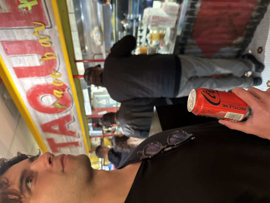 The image depicts the interior of Taquilla Taco Bar, located in Boston, United States. In the foreground, a male individual with dark, curly hair is seen in profile, looking towards the right side of the frame. He is wearing a black v-neck shirt, with a pair of dark-framed sunglasses hooked onto the collar. His right hand holds a red Coca-Cola Zero Sugar can, displaying a "RECYCLE ME" label.
In the midground, several other patrons are visible, standing in a line at a service counter. One man, seen from the back, wears a black puffer jacket and light grey pants, facing the counter. To his left, another man in a dark jacket is also facing the counter, and a woman with light brown hair is partially visible between them. Further left, another man in a dark hoodie is visible.
Above the counter, a large sign with a yellow border features the word "TAQUILLA" in prominent red letters, with "Taco bar" in smaller, yellow cursive letters below it. The serving counter below the sign displays various containers and a clear sneeze guard, suggesting food preparation or serving. The lower portion of the counter is constructed from corrugated metal, painted red. The floor is tiled with a white and brown geometric pattern.