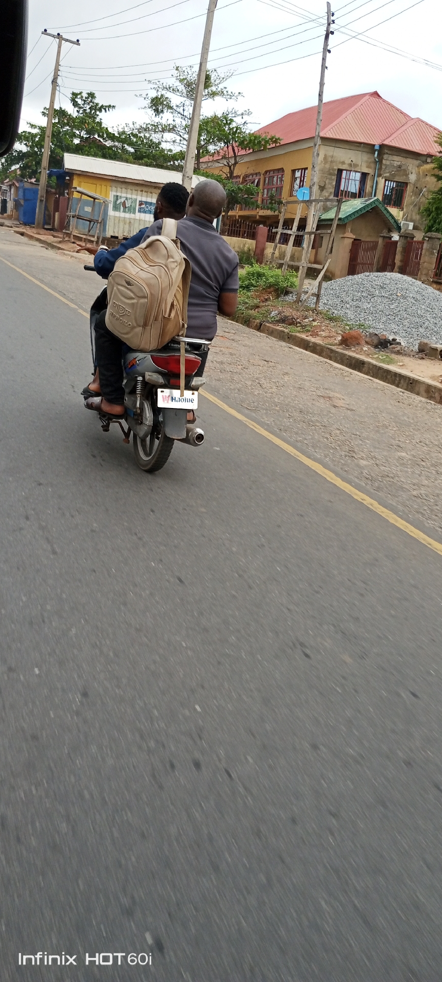 Two men are riding a motorcycle on a paved road in Adankolo, Nigeria. The motorcycle has a Haojue license plate and a tan backpack is strapped to the back of the seat. One man is wearing a gray shirt, and the other is wearing a blue jacket and sandals. The road has a yellow line marking the lane. Buildings, utility poles, and piles of gravel are visible on the roadside. The photo was taken with an Infinix HOT 60i phone.