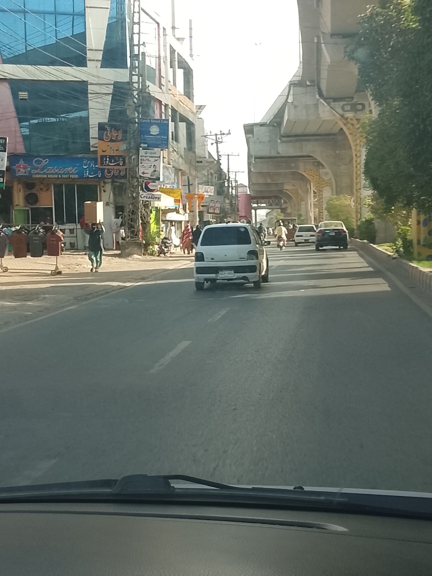 The image captures a street view, presumably taken from inside a moving vehicle, with a road stretching into the distance. A light-colored vehicle is prominently visible ahead, along with other cars and motorcycles in the distance. Pedestrians are seen on the sidewalks alongside the road, and a man carries a box on his head. The presence of commercial establishments, including "Lasania Chaargha House & Fast Food" and "Creators," indicates an urban setting. An elevated structure, possibly a flyover or metro line, looms over the road. The scene is bathed in sunlight, and the overall atmosphere seems bustling with activity.