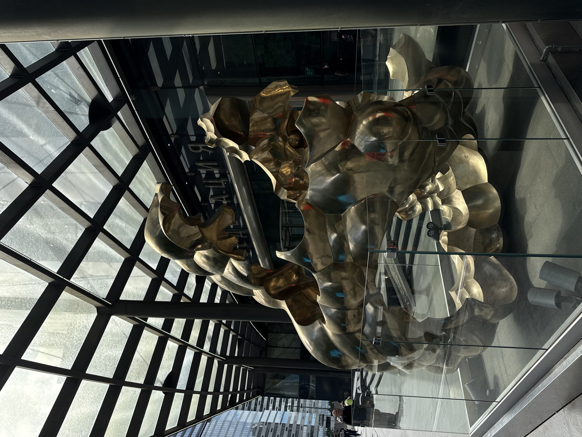 Captured from a low-angle perspective, the image features a golden abstract sculpture displayed in a glass enclosure within a modern building. Sunlight streams through a grid-patterned glass roof, casting reflections and highlights on the sculpture's surface. The sculpture, comprised of bulbous, organic forms, dominates the scene. In the reflection visible in the glass, faint figures can be discerned, appearing to be people walking outside. The building's exterior and cityscape stretch out in the background, indicating an urban setting. Based on information available, this is potentially in Khimki, Russia.