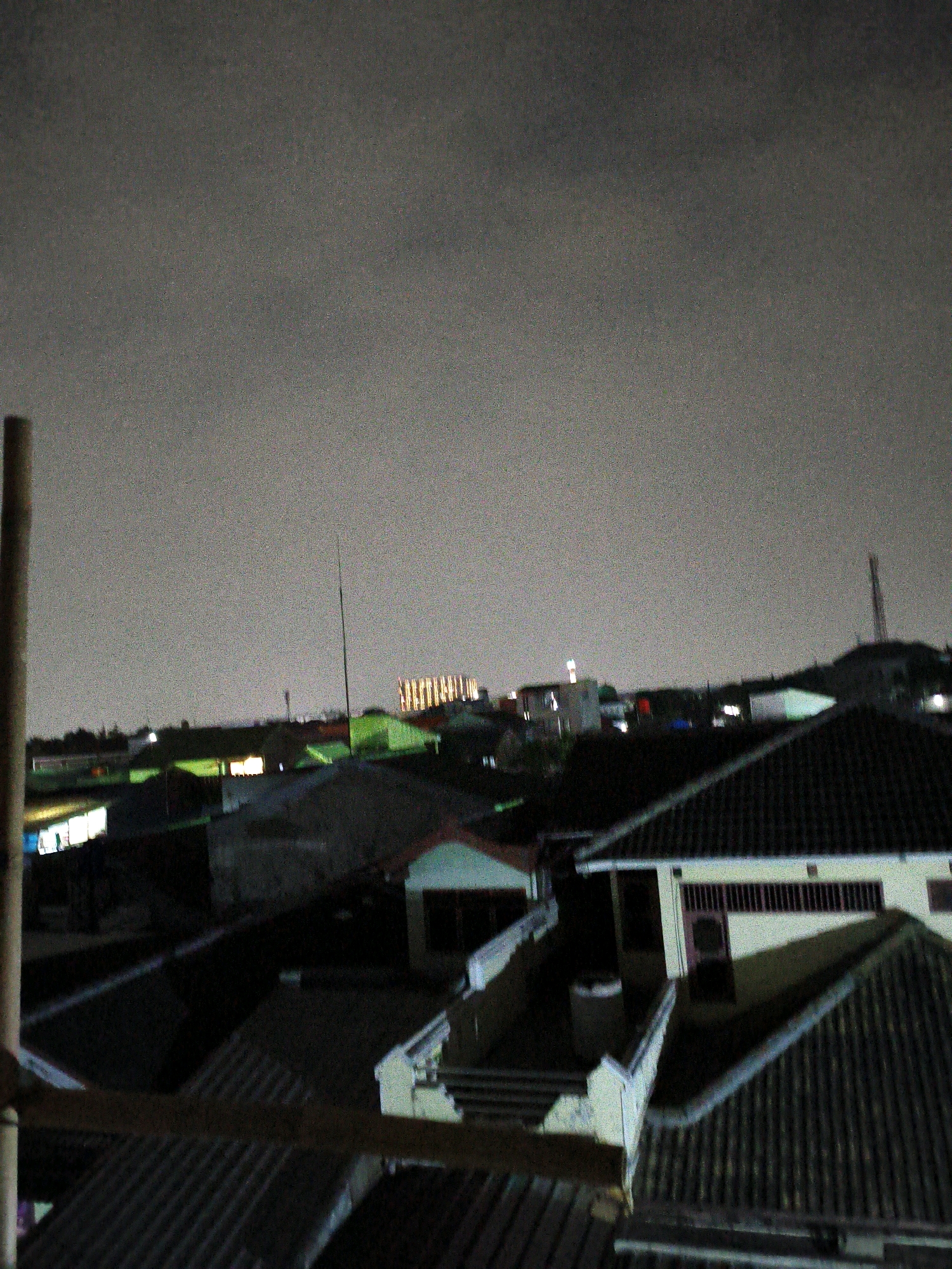 This is a wide-angle, aerial nighttime photograph captured from an elevated vantage point, likely a rooftop or balcony, overlooking a dense urban residential area in Bekasi, Indonesia. The scene is dominated by the muted, diffused light of the night sky, creating a hazy and somewhat grainy atmosphere.
The foreground is occupied by the angled roofs of several houses, constructed with dark, tiled materials. A rough, vertical wooden pole or structure is visible on the far left, partially obscuring the view and suggesting the perspective is from a constructed elevated area.
In the mid-ground, more rooftops are visible, along with the outlines of buildings and structures. Some windows emit a warm, artificial light, indicating occupancy within the residences. Green-tinged lighting emanates from some structures, possibly due to streetlights or specific architectural lighting. A tall, slender antenna or pole stands prominently in the mid-ground.
The background reveals more distant buildings and infrastructure. A large, brightly lit building with a grid-like pattern of windows is visible in the far distance, standing out against the darker urban landscape. Other faint lights and structures dot the horizon. The overall impression is of a densely populated suburban or semi-urban environment at night, characterized by consistent building density and varying levels of artificial illumination. The lack of visible people or distinct activity points to a quiet, observed moment in time.