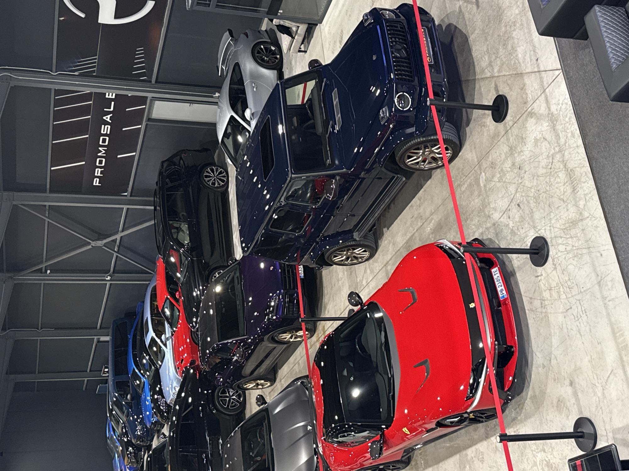 An indoor automobile showroom displays a lineup of luxury vehicles. The cars, varying in colors of blue, silver, black, purple, and red, are neatly aligned. A red Ferrari and a dark blue G-Wagon Mercedes stand out in the foreground. "PROMOSALE" is visible in large letters along the ceiling of the showroom, which is illuminated by overhead lighting. The location is Plovdiv, Bulgaria. The cars are placed behind red ribbon barriers supported by posts with circular bases.